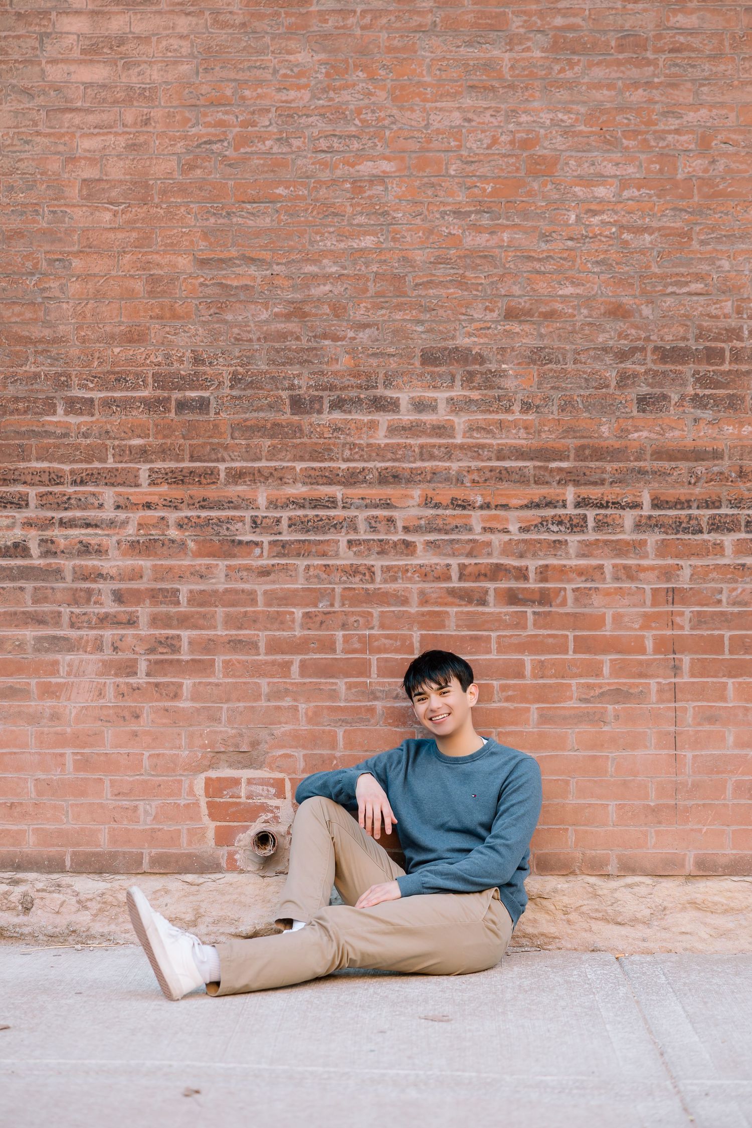 Casual portrait against weathered brick wall wearing blue sweater and beige pants.