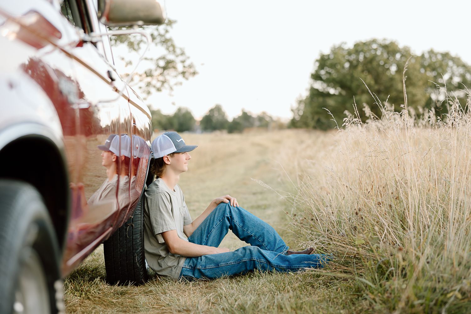 Someone sitting beside a red pickup truck in tall grass during golden hour.