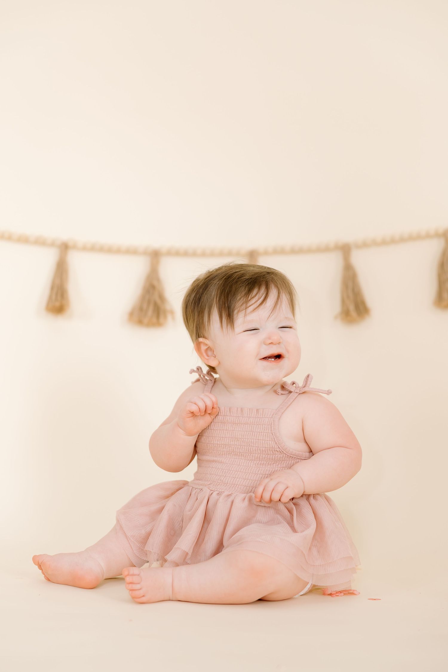 Smiling baby in pink dress sits beneath decorative tassel garland on cream background.