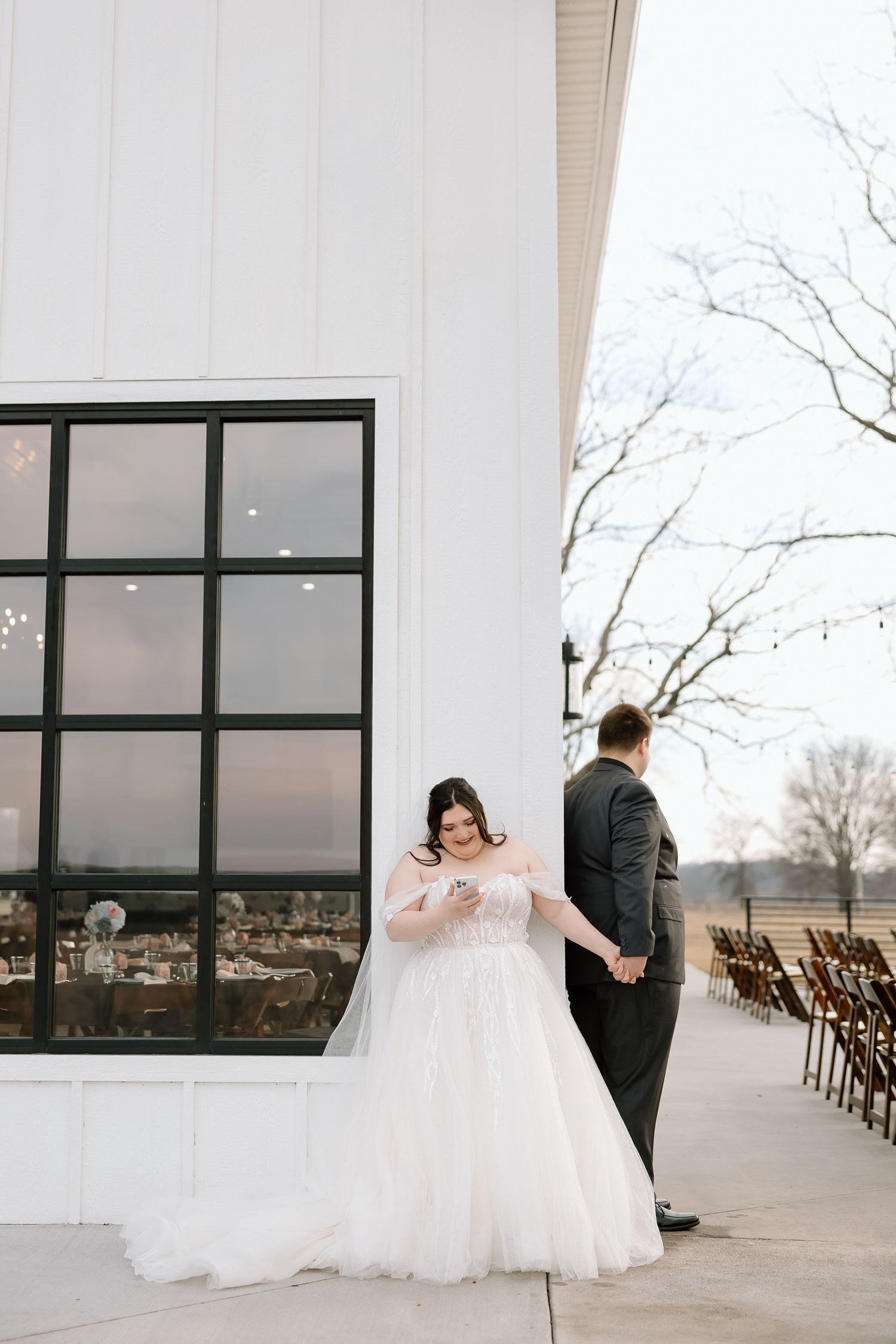 Bride checking her phone while groom stands separately during wedding photoshoot.