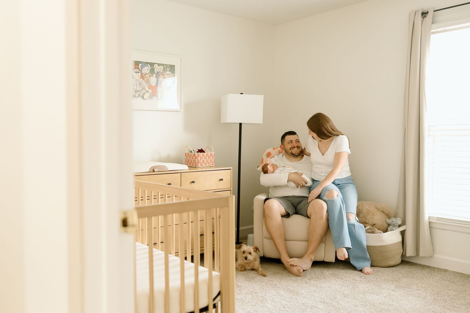 Parents sitting in nursery with baby next to wooden crib and floor lamp.