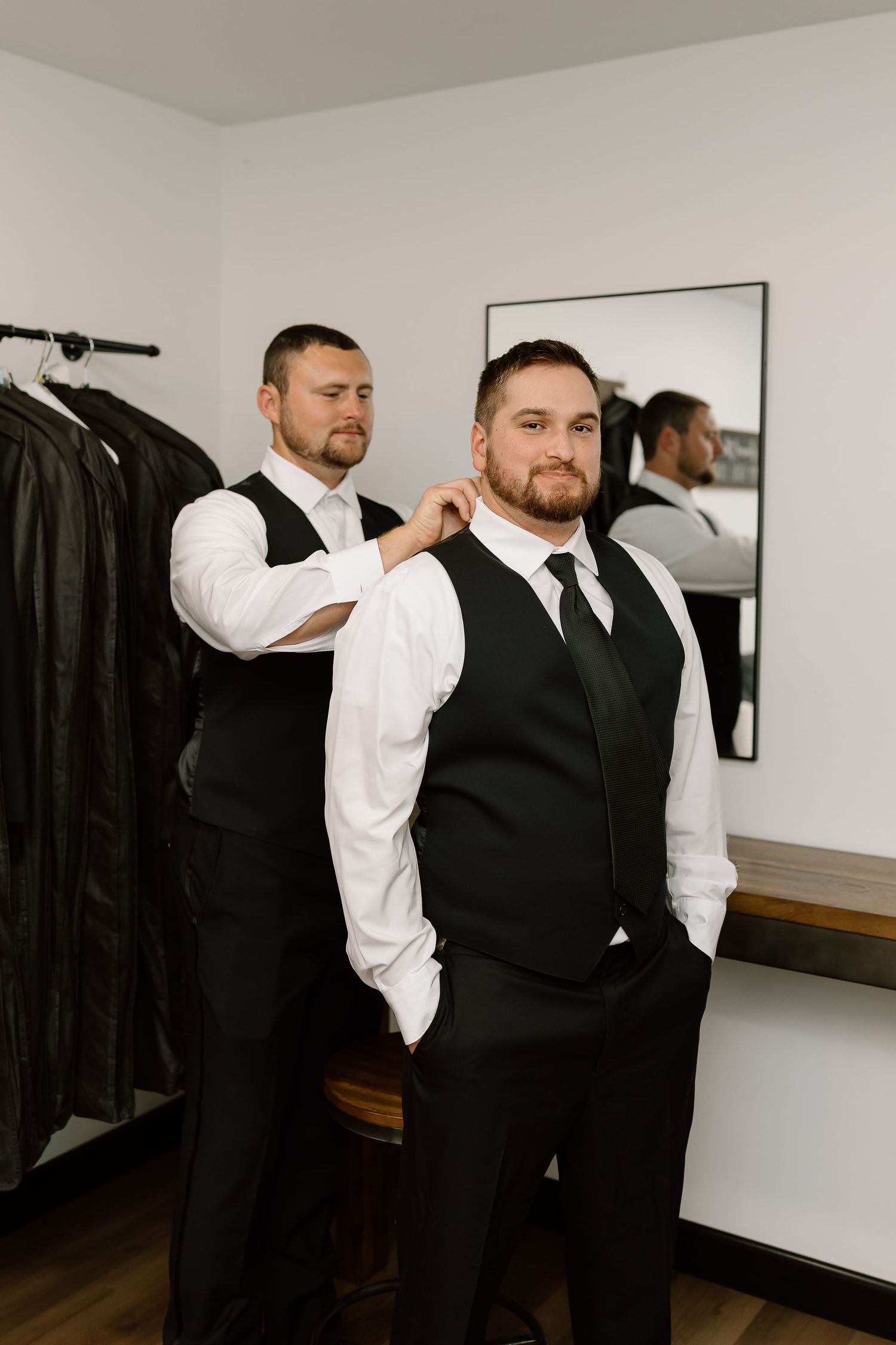 Two people in formal attire getting dressed in a room with a mirror and dark clothing hanging on the wall.