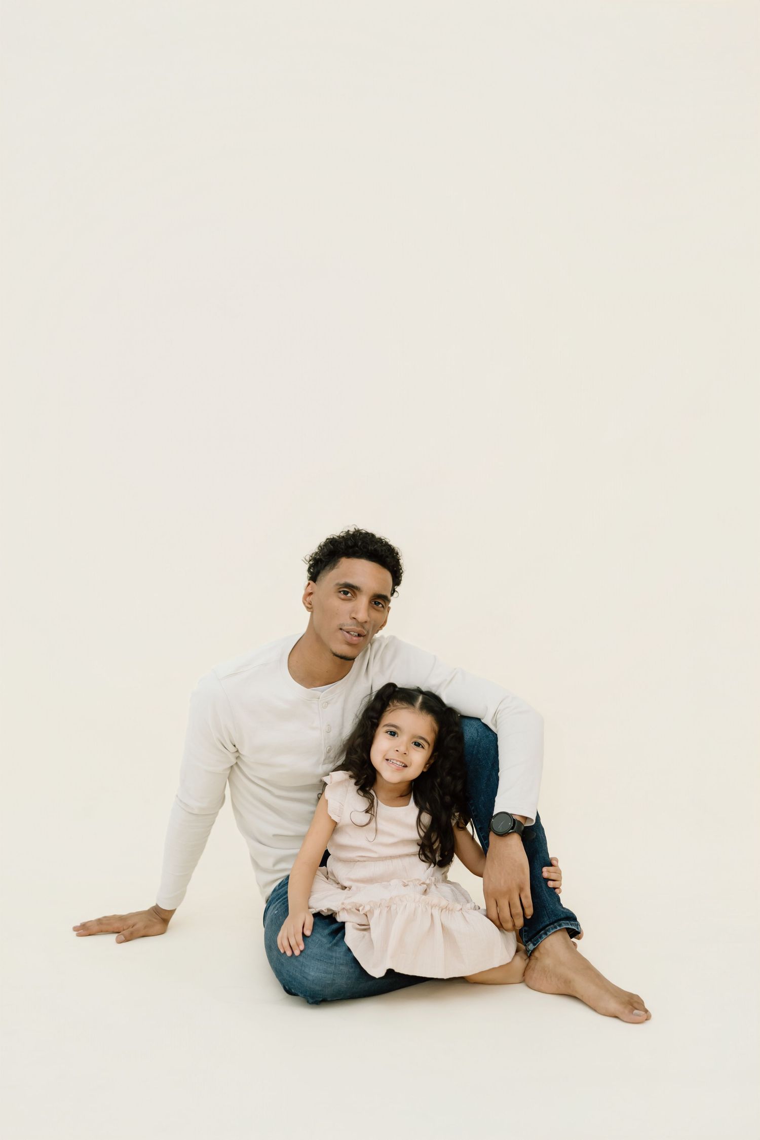 A parent and child sit together on a white backdrop wearing light colored clothing for a minimalist portrait.