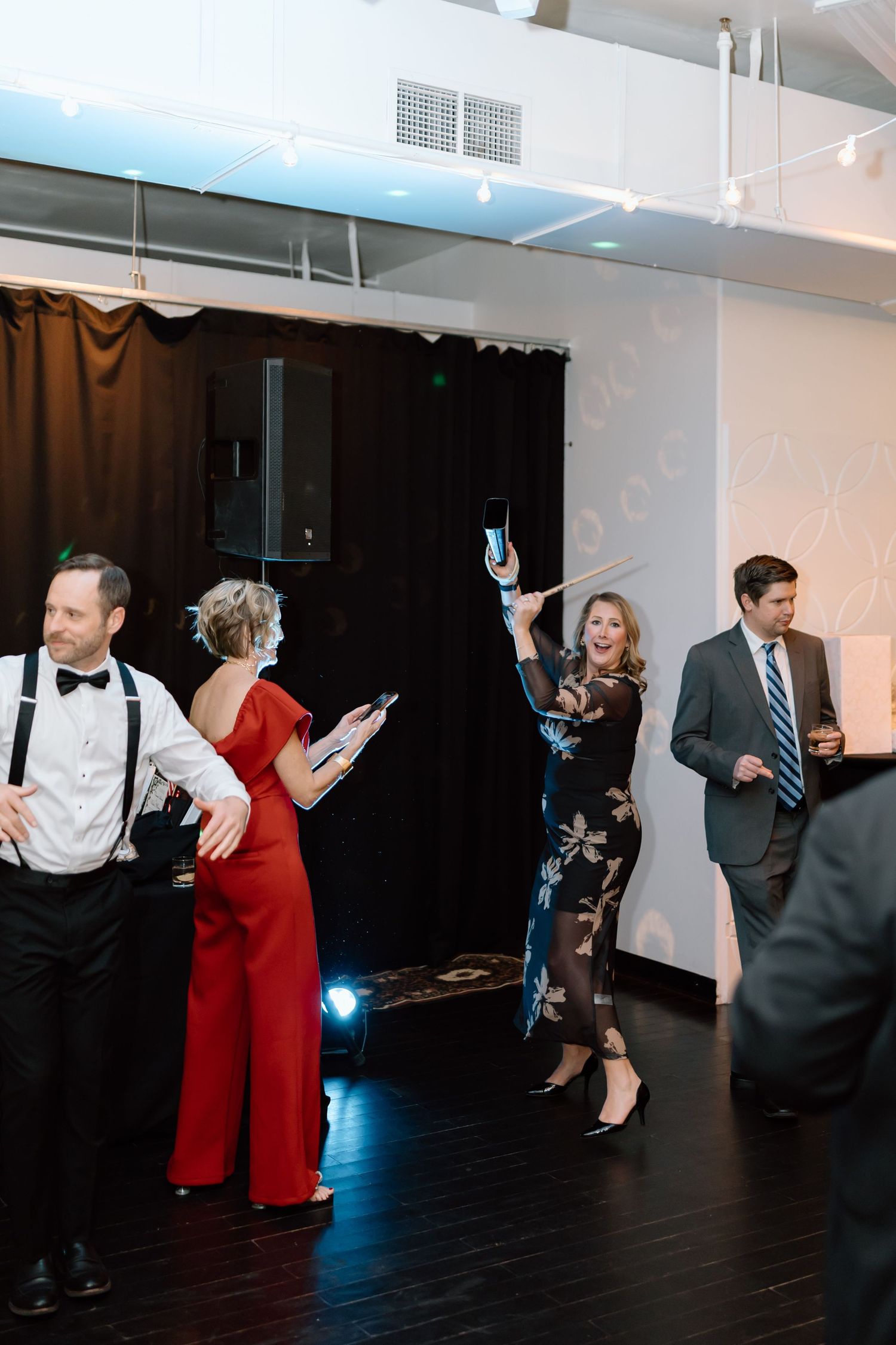Party guests in formal attire dancing and celebrating at an indoor event with dark hardwood floors and white walls.