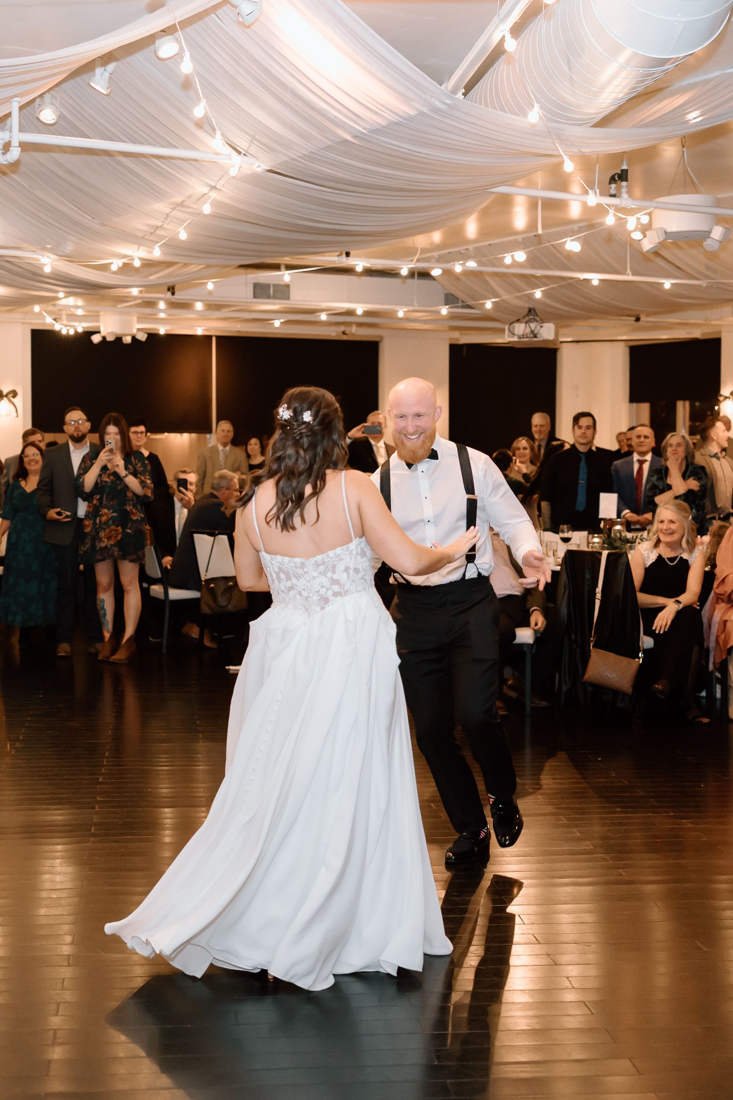 A couple shares their first dance at a wedding reception under draped fabric and string lights on a dark wooden dance floor.