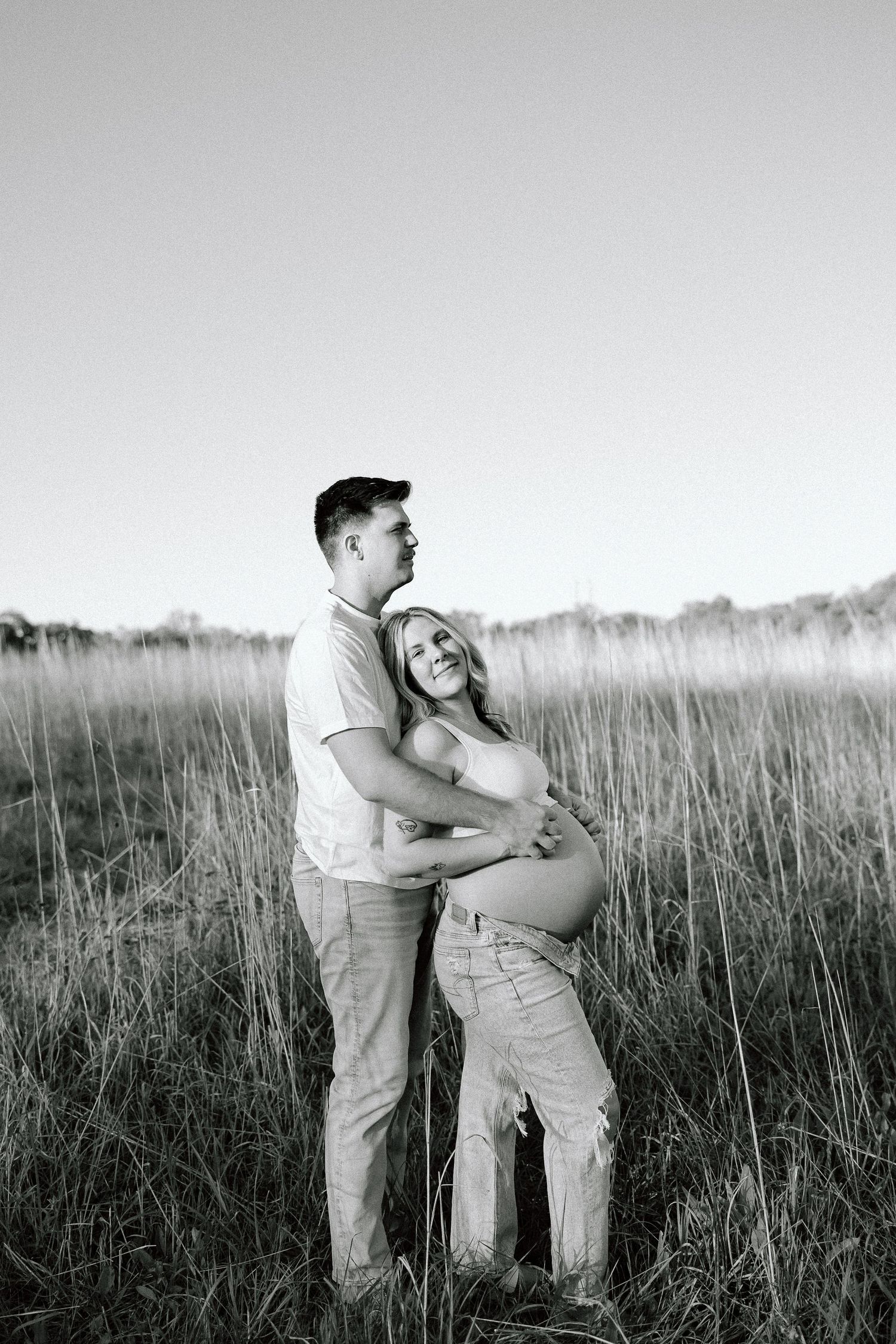 Black and white photos show a couple embracing in a field of tall grass against a bright sky.