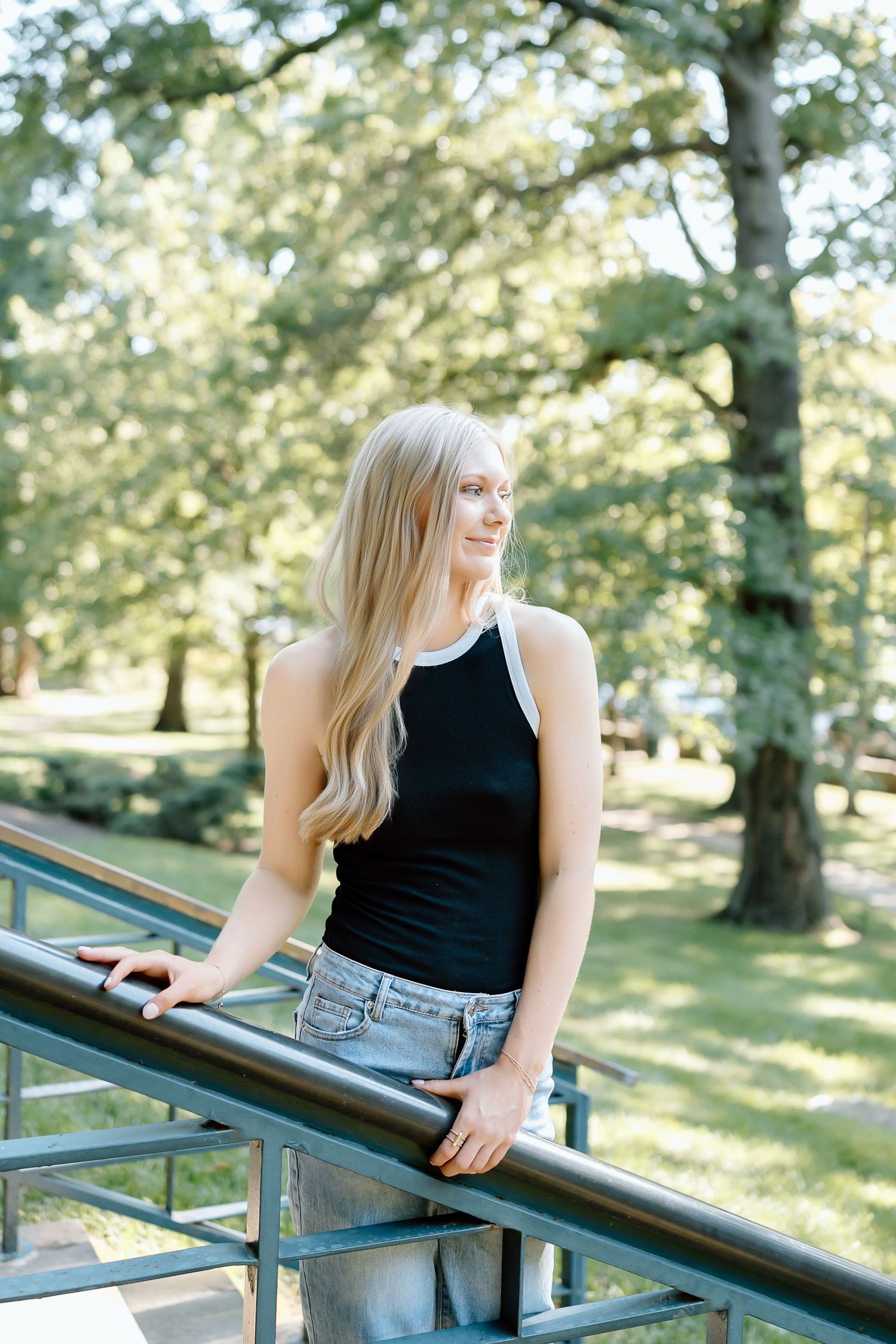 Person in black tank top and jeans sits casually on metal railing in sunny park setting.