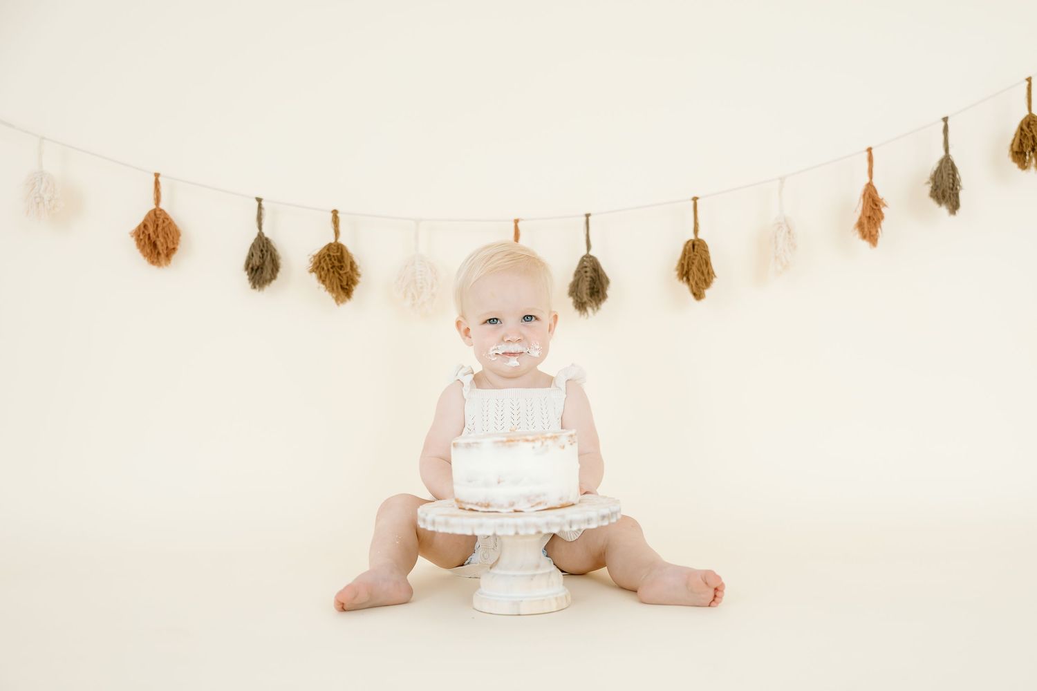 A baby in a white dress sits on the floor while holding a birthday cake with pink frosting and candles.