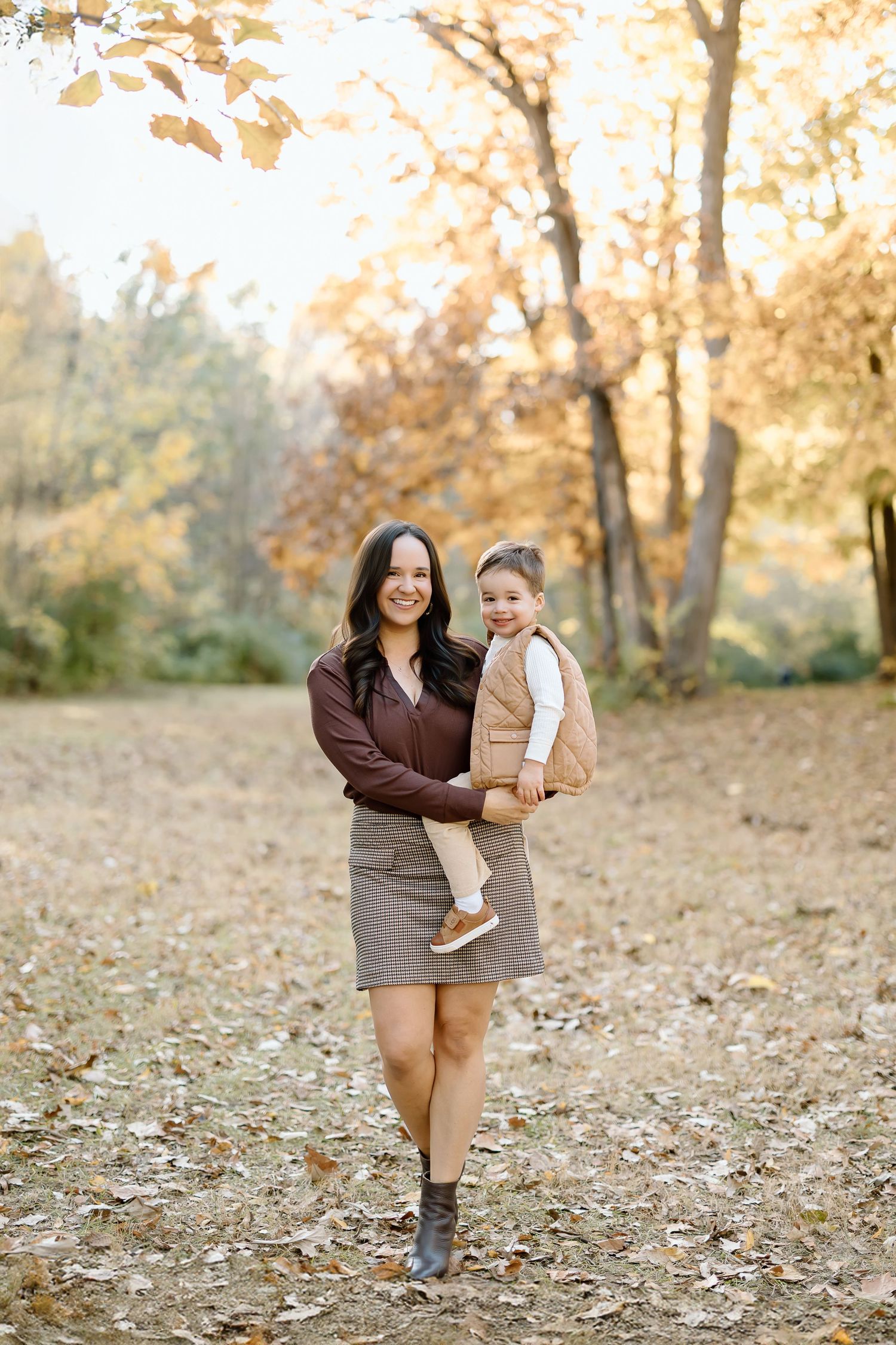 A mother and child share tender moments while walking through fallen autumn leaves in a park setting.
