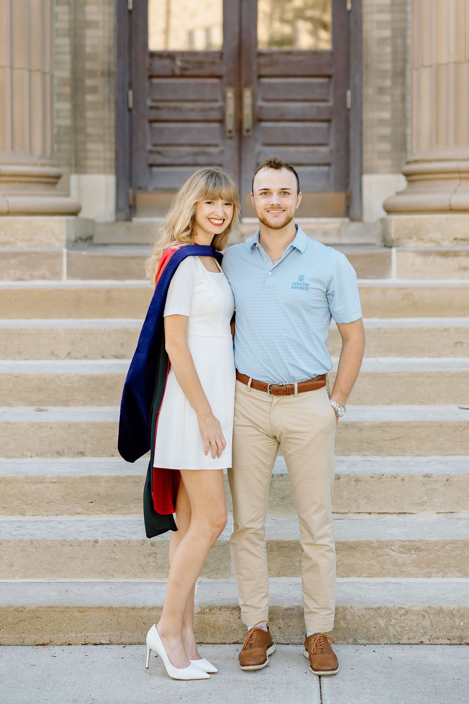 A couple in casual attire poses together on stone steps outside a building with elegant wooden doors.