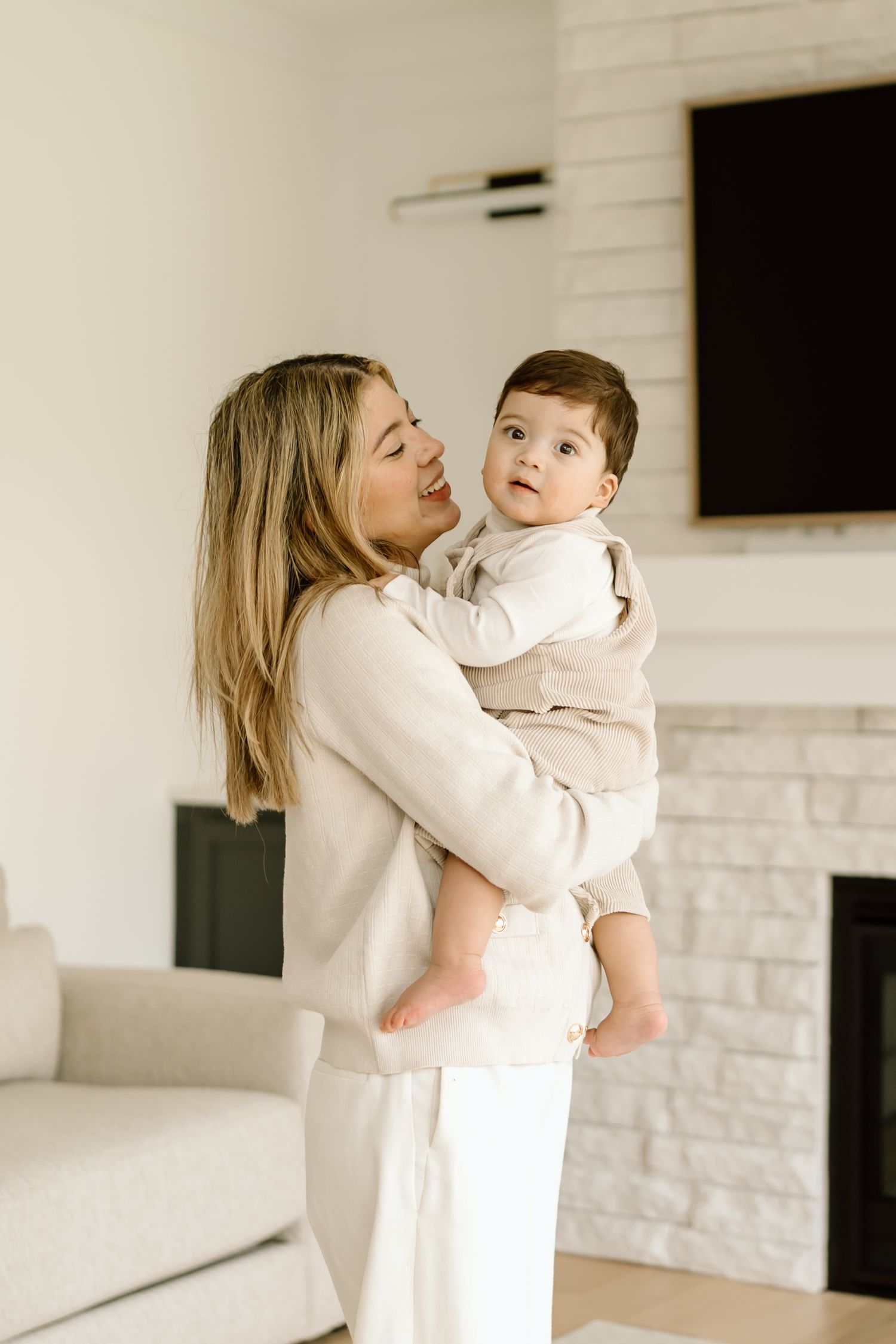 Parent in white sweater holds and lovingly interacts with baby against white brick wall background.