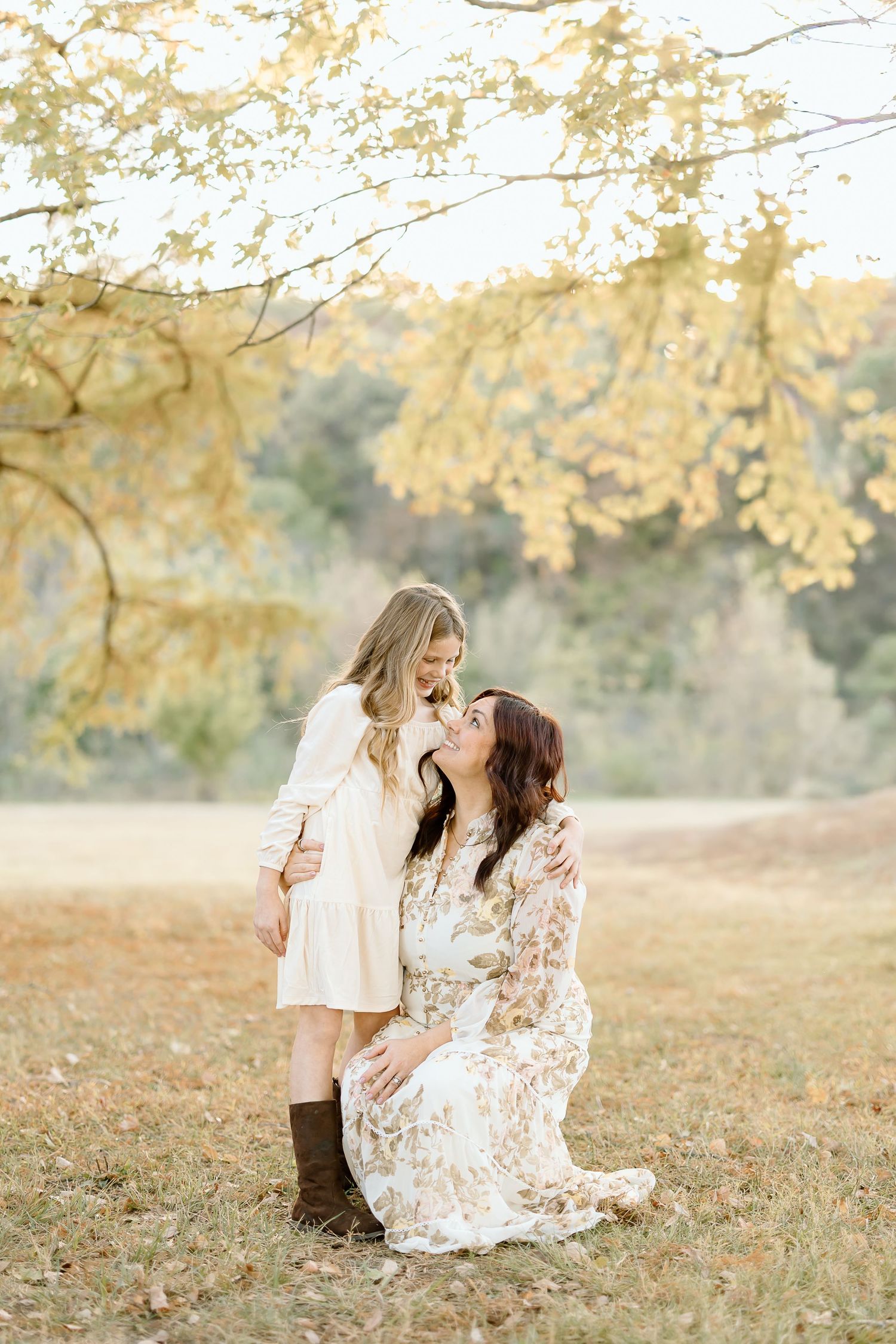 Two people in light floral dresses share a tender moment under golden autumn trees in a sunlit field.