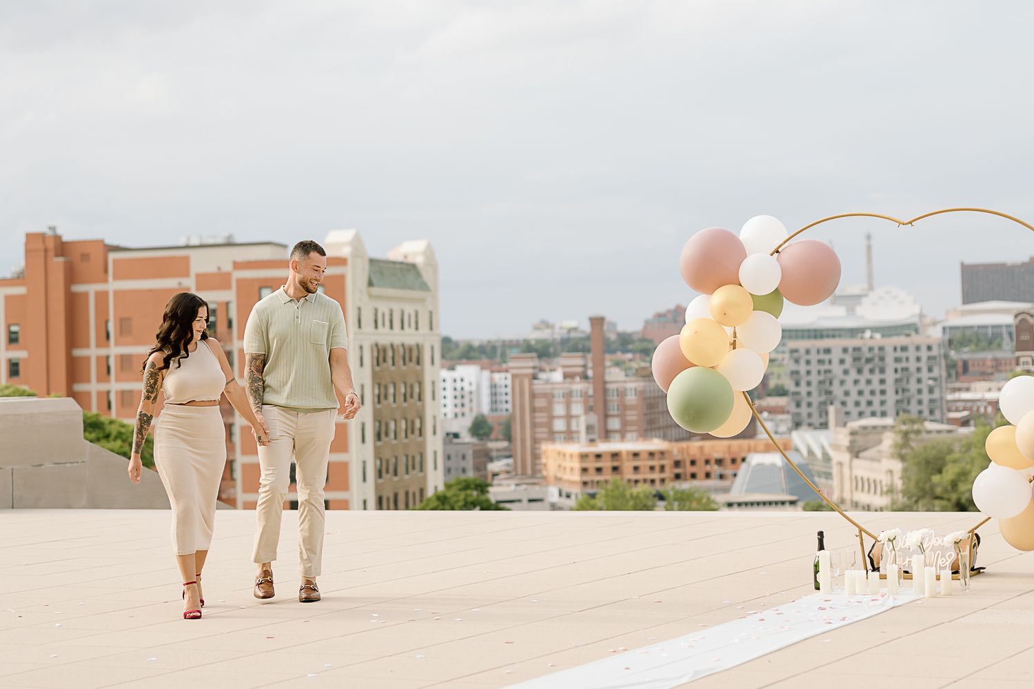 Couple walking on rooftop with colorful balloons and city skyline in background.