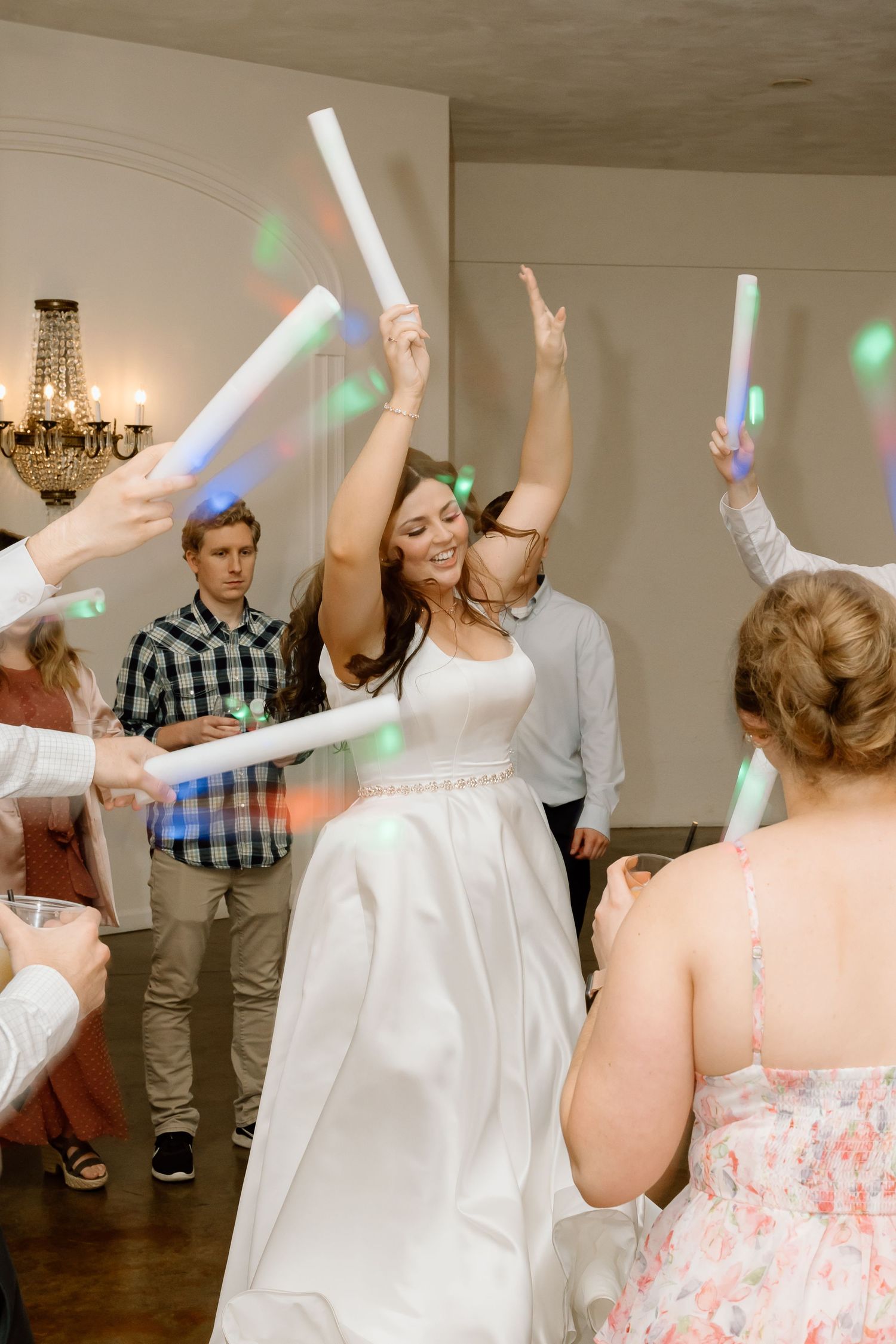 People dancing and waving light sticks at a wedding reception celebration with disco lights and chandeliers.