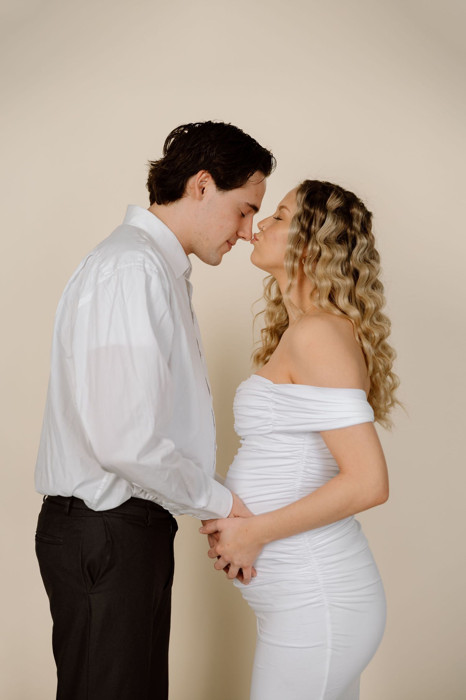 A romantic maternity photo series showing a couple in formal attire embracing against a neutral backdrop.
