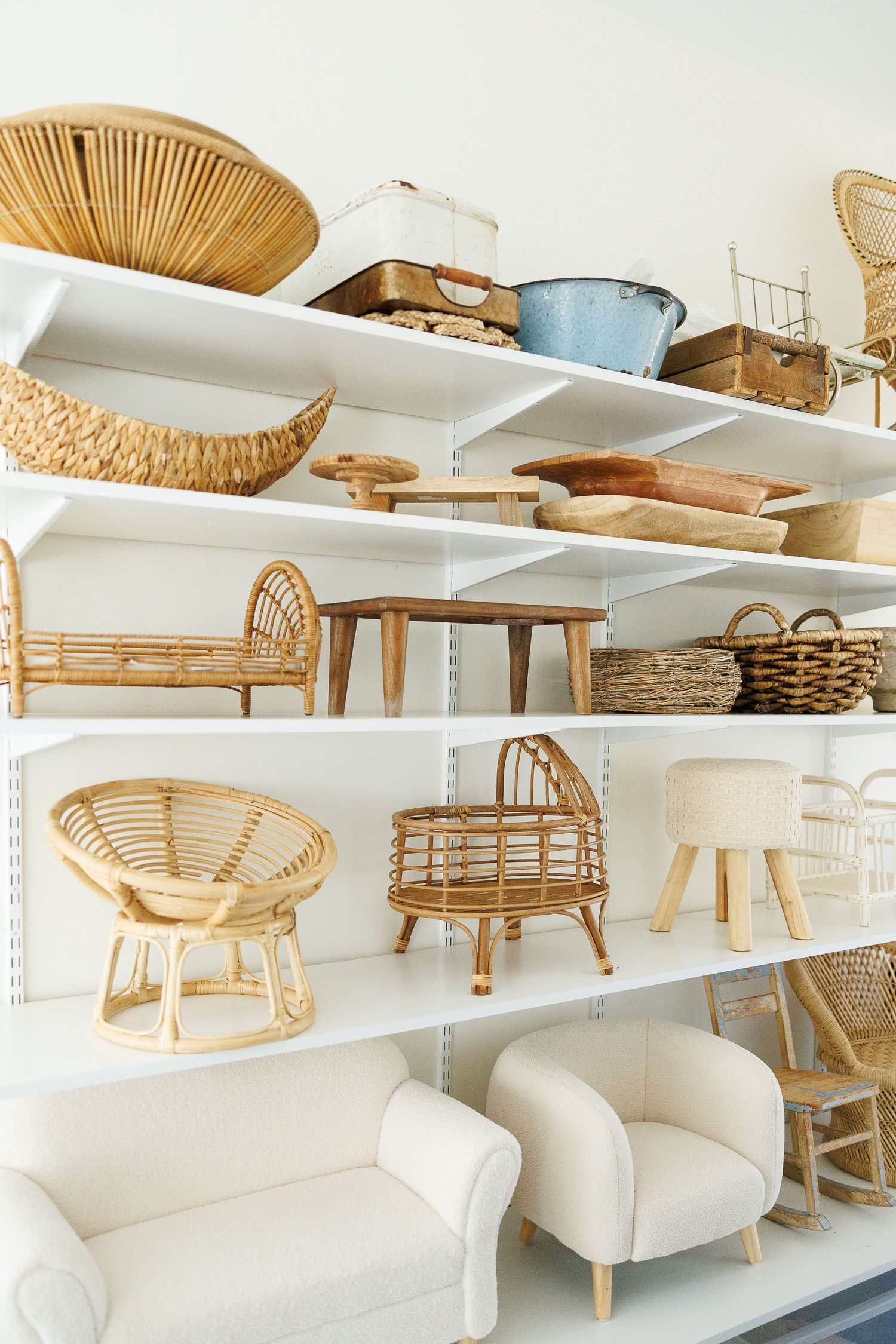 Modern white shelving unit displaying rattan furniture, wicker baskets, and cream upholstered seating in a bright minimal space.
