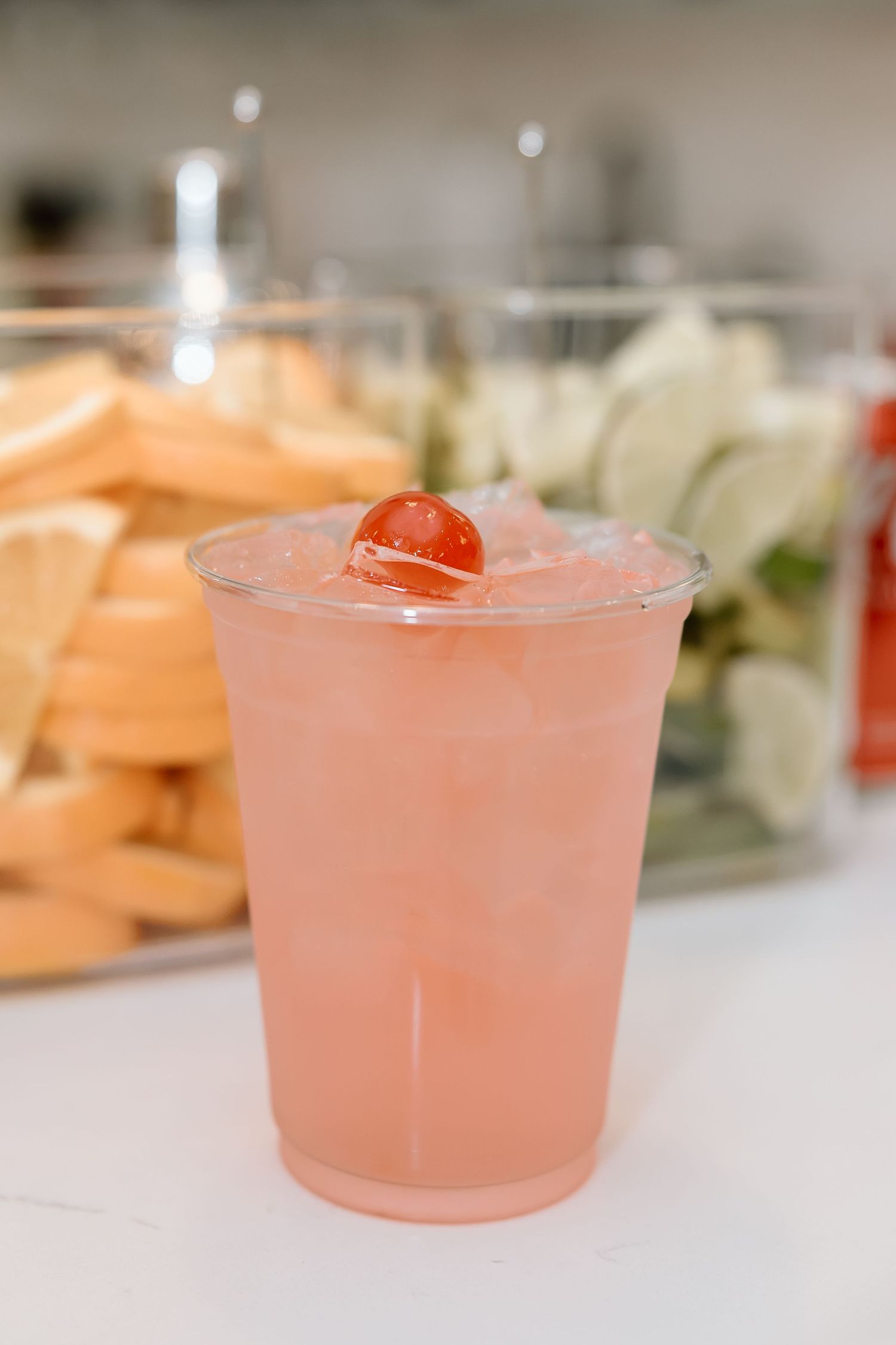 A pink cocktail in a clear plastic cup garnished with a maraschino cherry, with appetizers visible in the background.