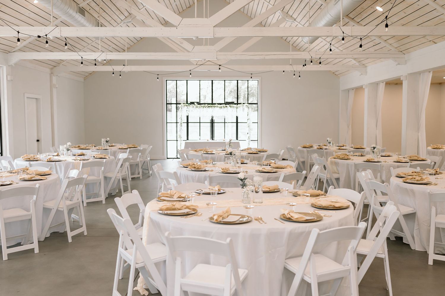 Modern white wedding reception venue with round tables, white linens, and gold charger plates.