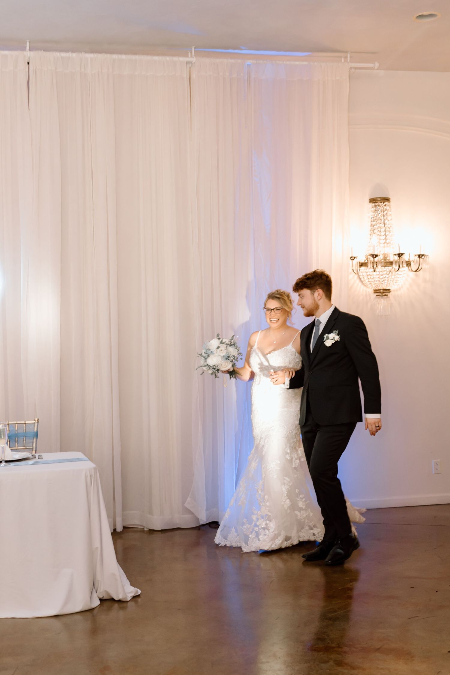 A couple enters their wedding reception venue with white drapes and a crystal chandelier in an elegant indoor setting.
