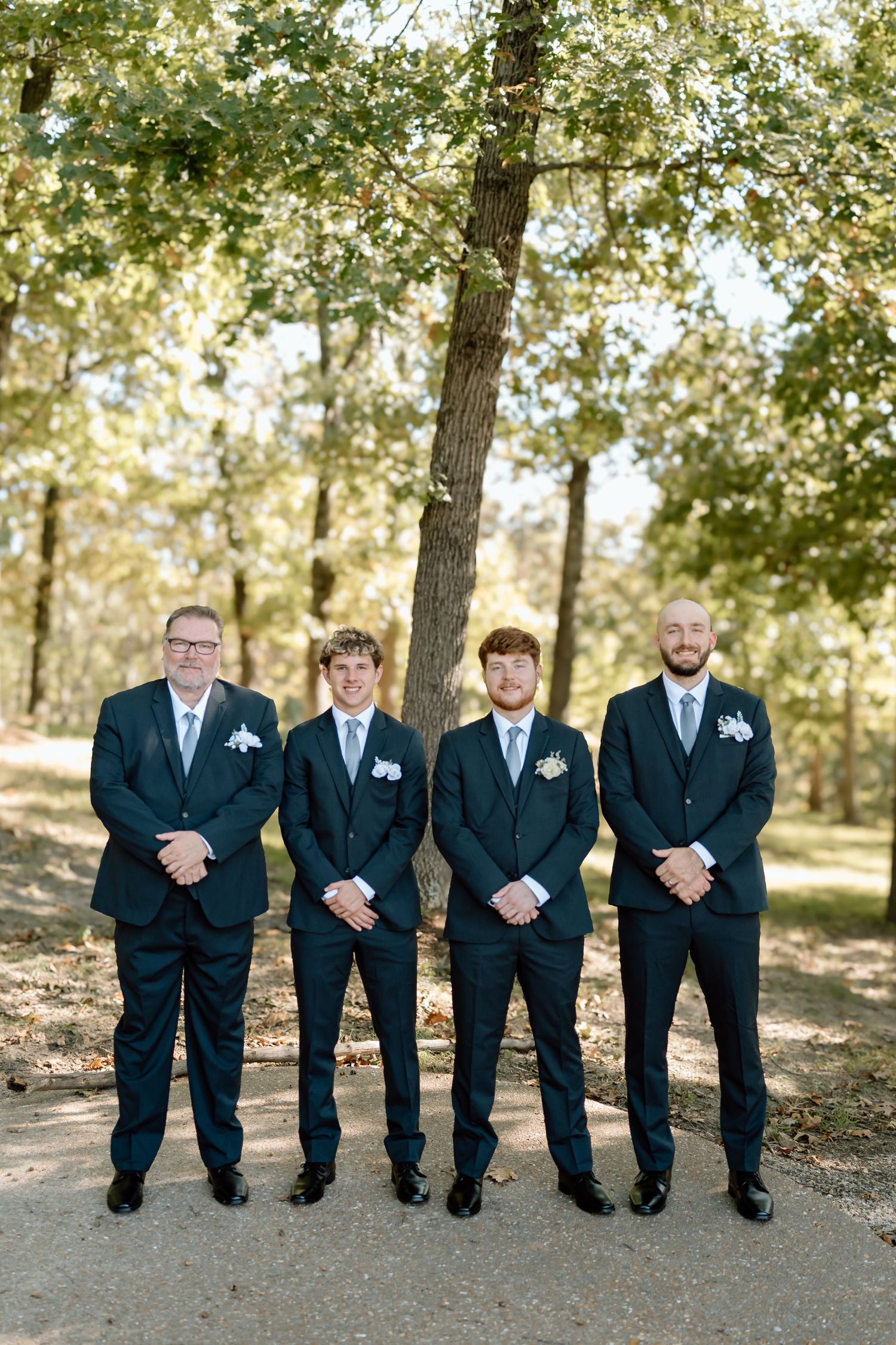Groomsmen in matching black suits and ties stand together outdoors in a wooded area for wedding photos.