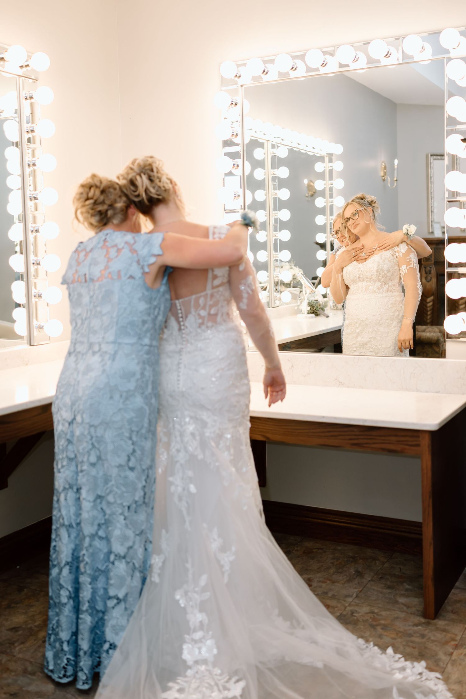 Two people share an emotional embrace in a dressing room with illuminated makeup mirrors and white decor.