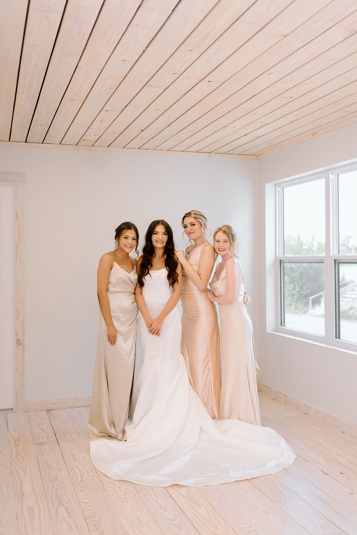 A bridal party in blush and white dresses shares a joyful moment together in a bright white room.