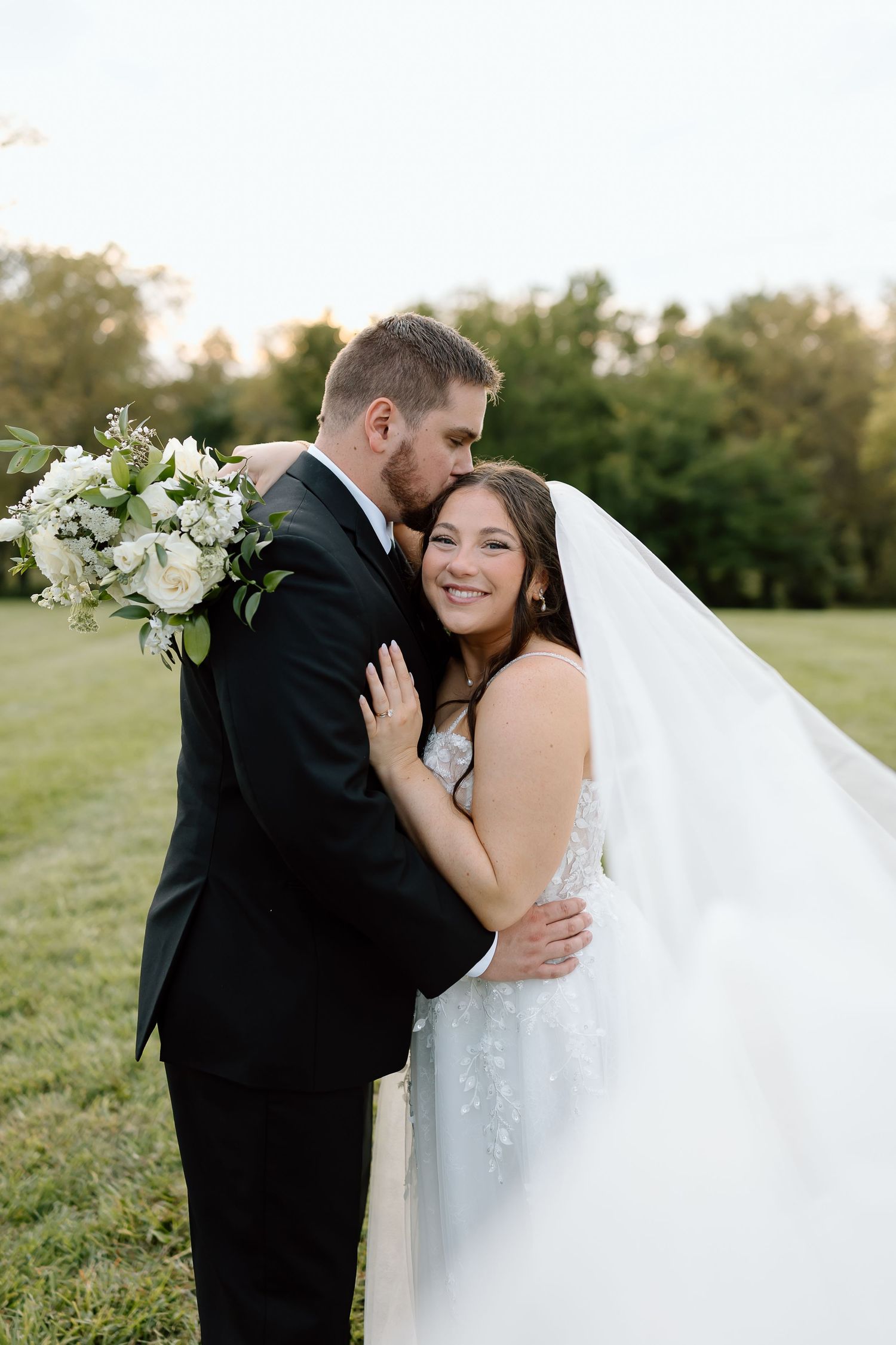 A romantic wedding couple embraces outdoors in a grassy field during golden hour.