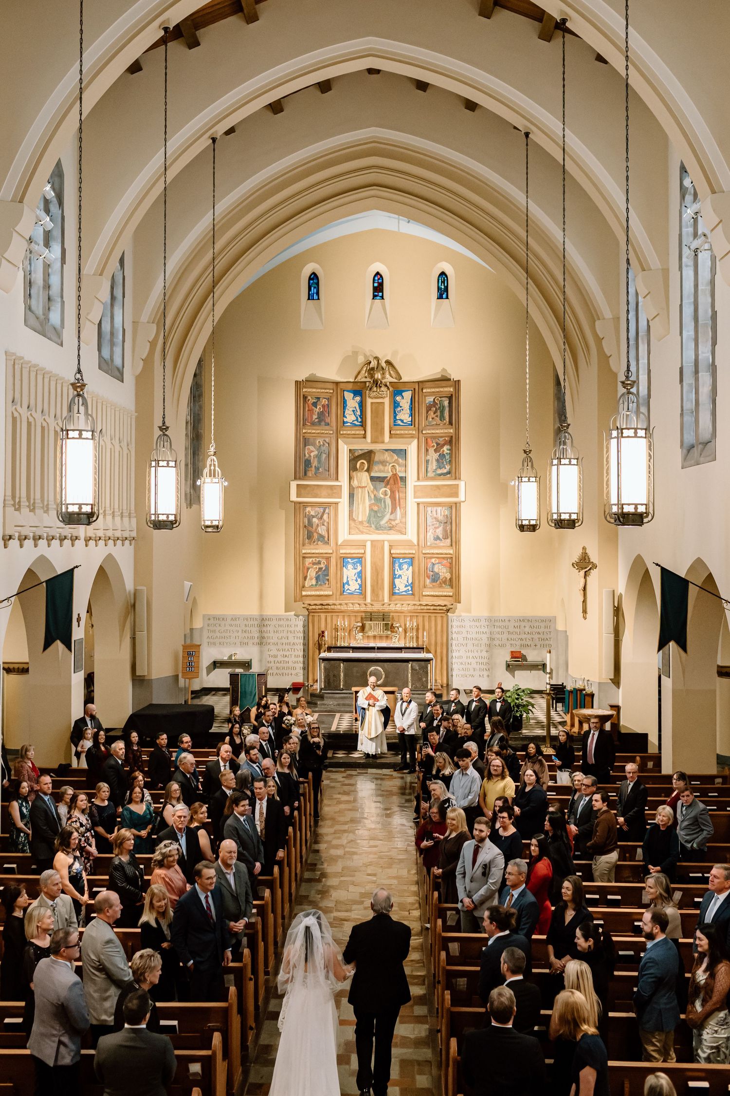 A wedding ceremony takes place inside a grand cathedral with arched ceilings, hanging lanterns and pews filled with guests.