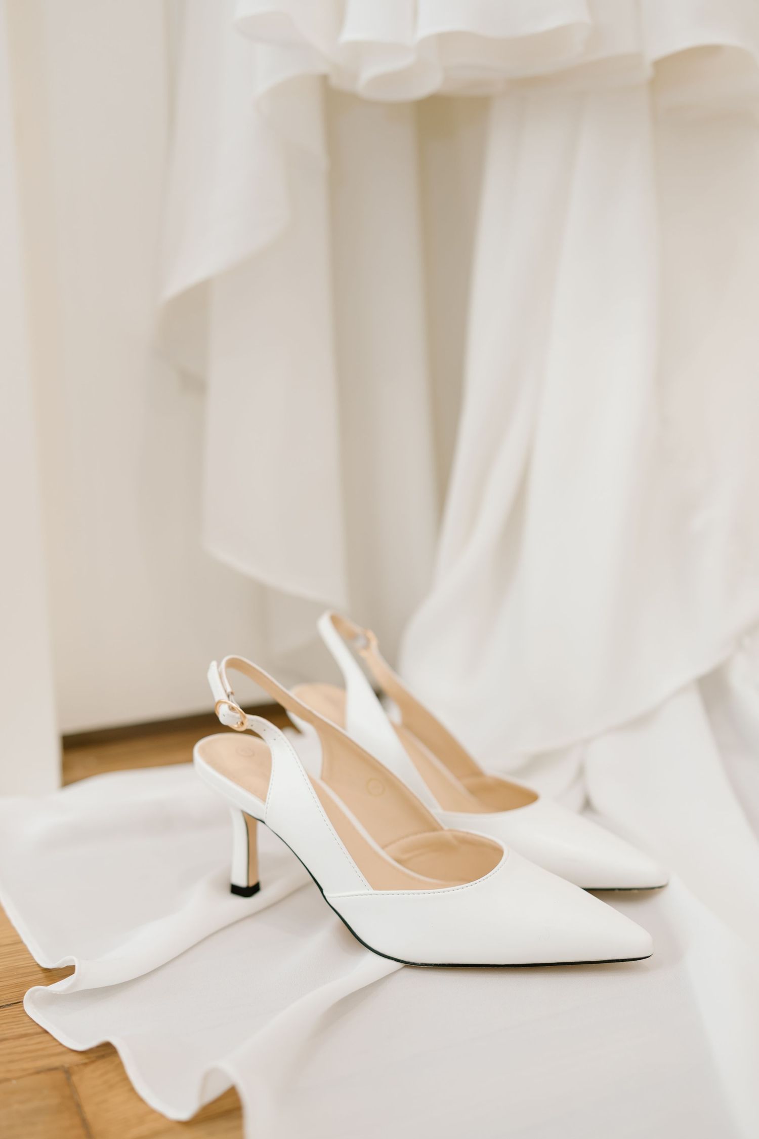 Elegant white bridal shoes with pointed toes and kitten heels sit beside flowing white fabric on a wooden floor.