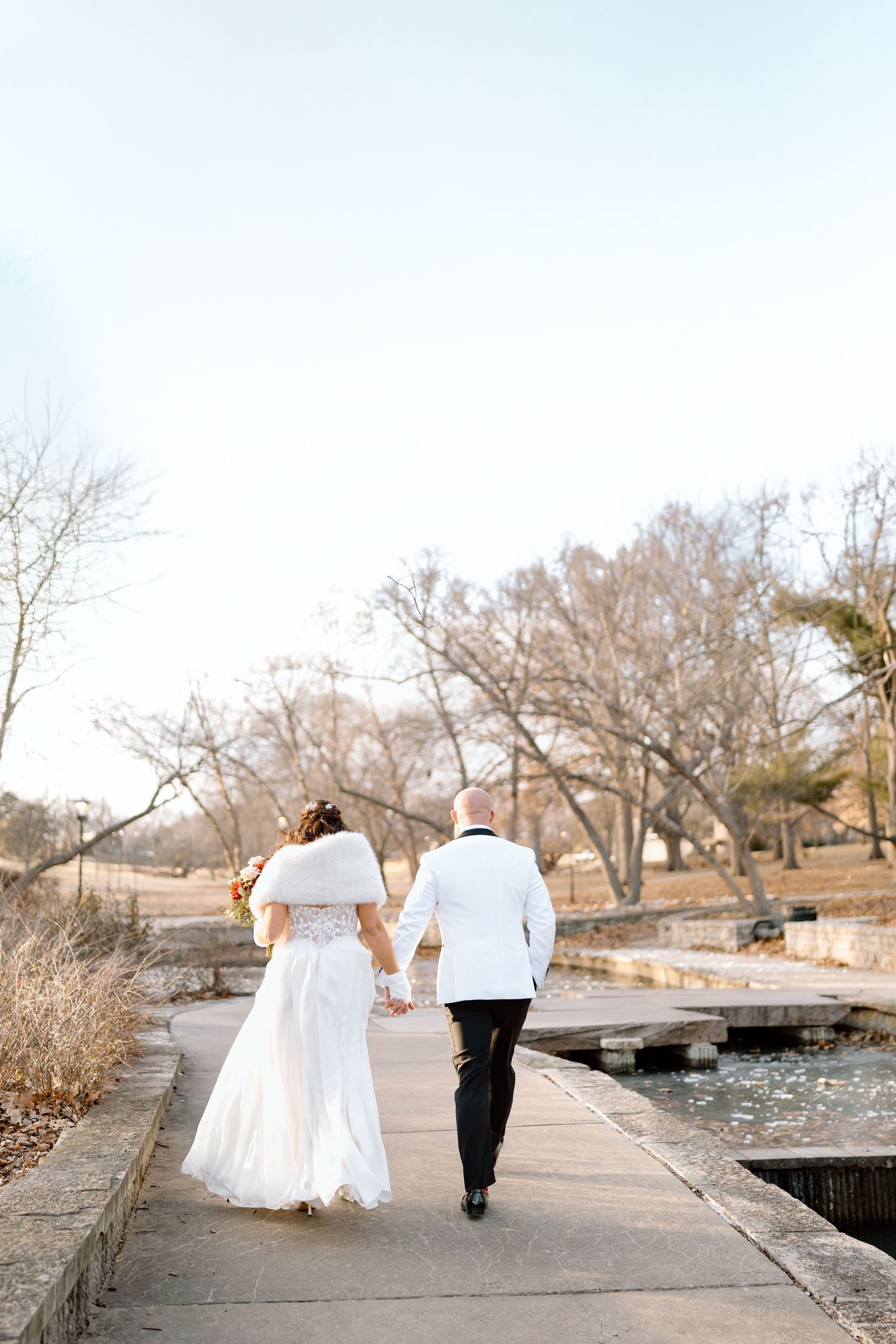A newlywed couple walks hand in hand along a lakeside path on their wedding day.
