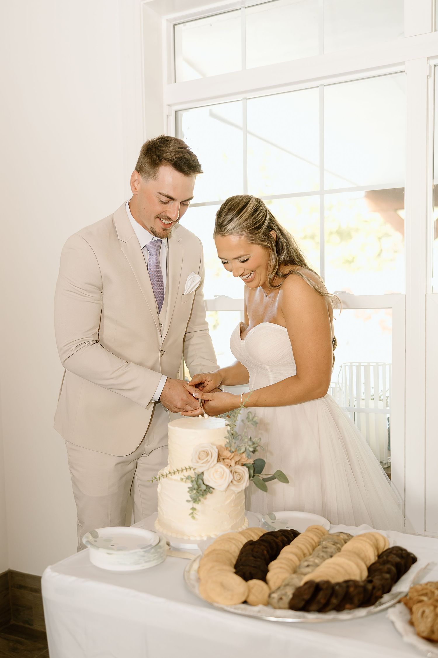 Couple cuts their elegant white wedding cake decorated with eucalyptus at reception.