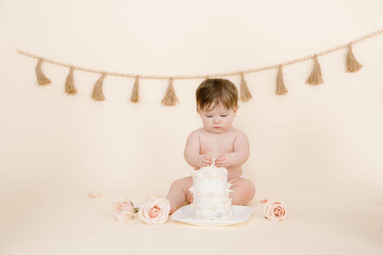 A baby sits with a white cake decorated with roses and a tassel banner backdrop.