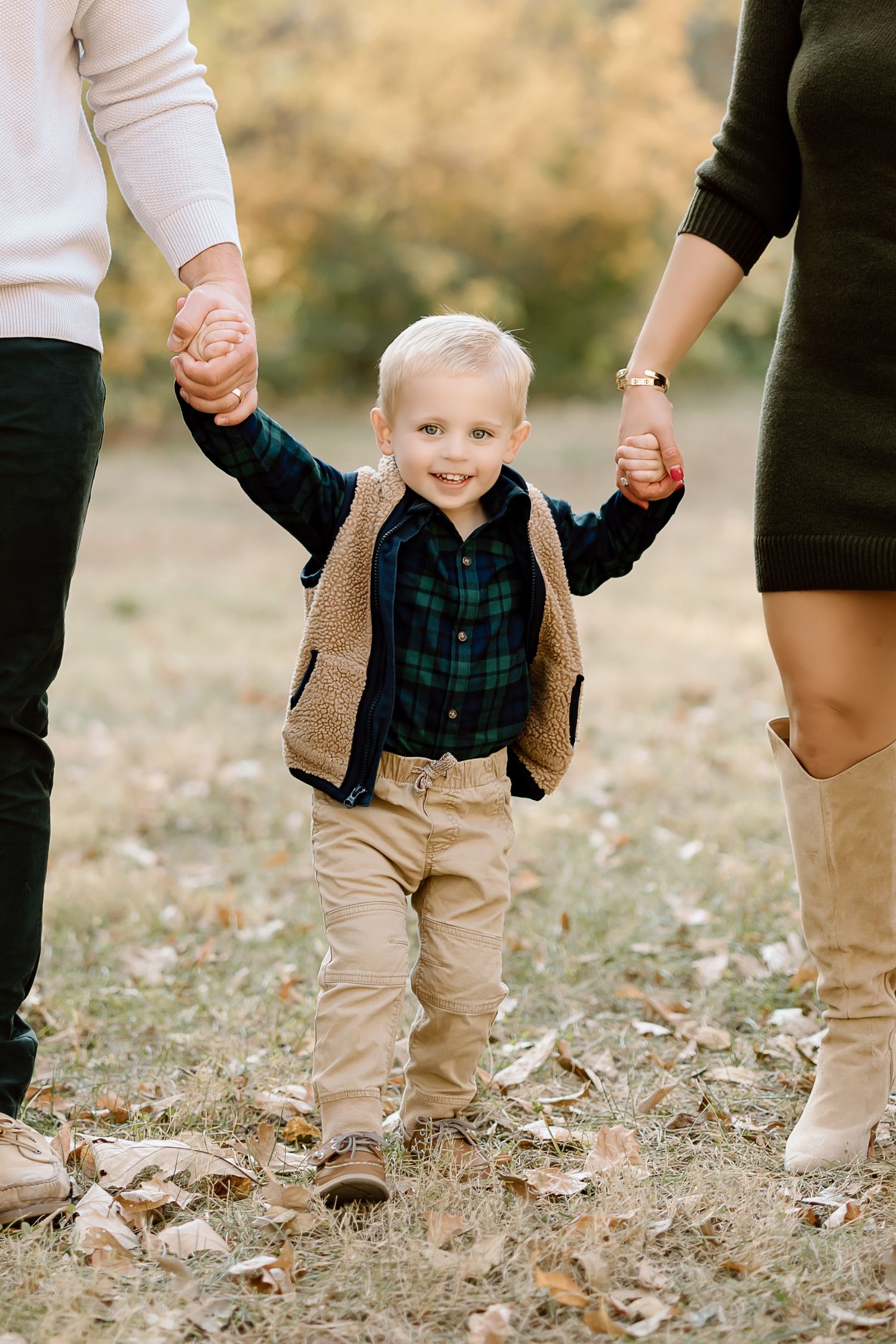 Young child in plaid shirt and vest holds hands with parents while standing in fallen leaves.