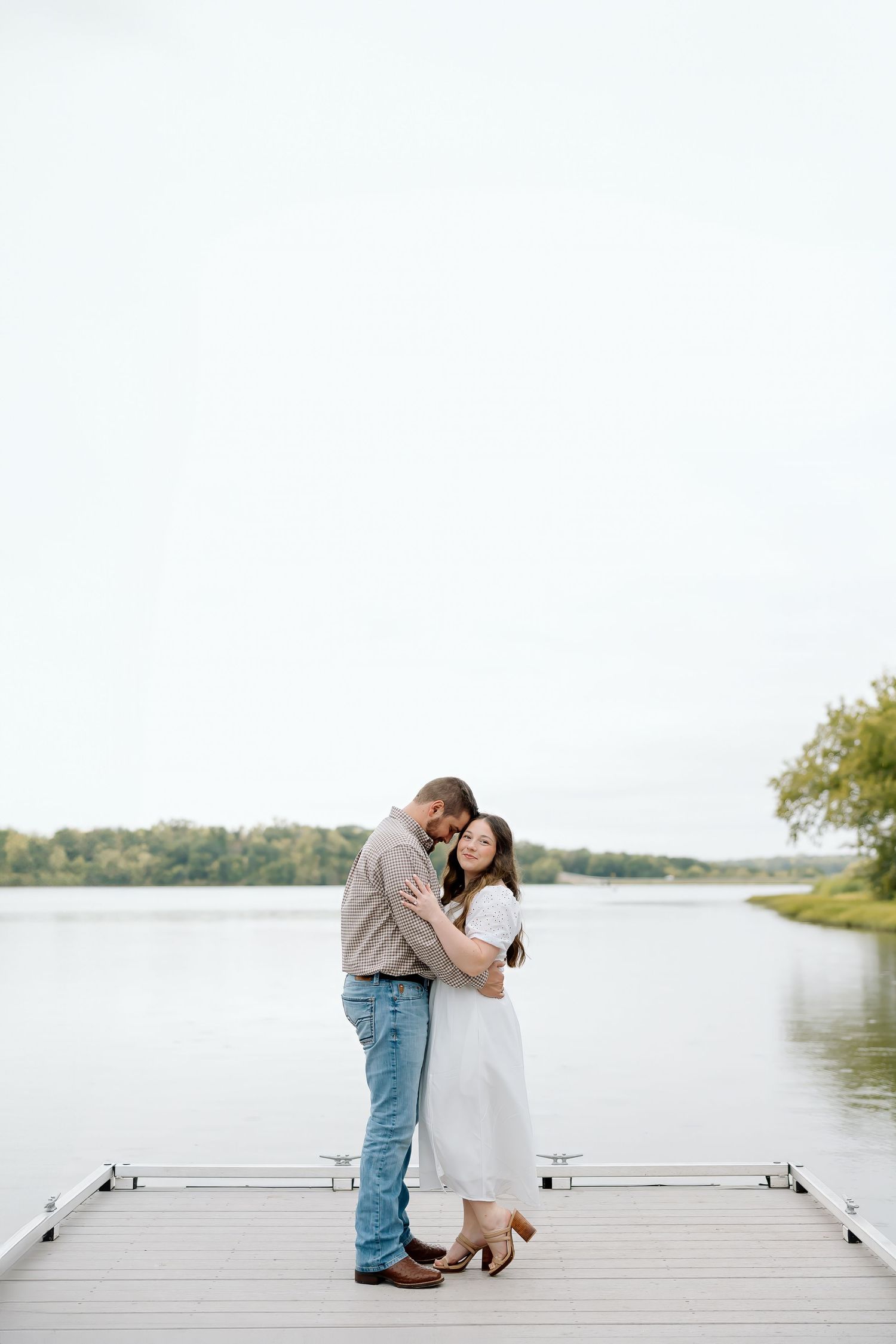 A couple shares a tender embrace while standing on a wooden dock over calm waters.