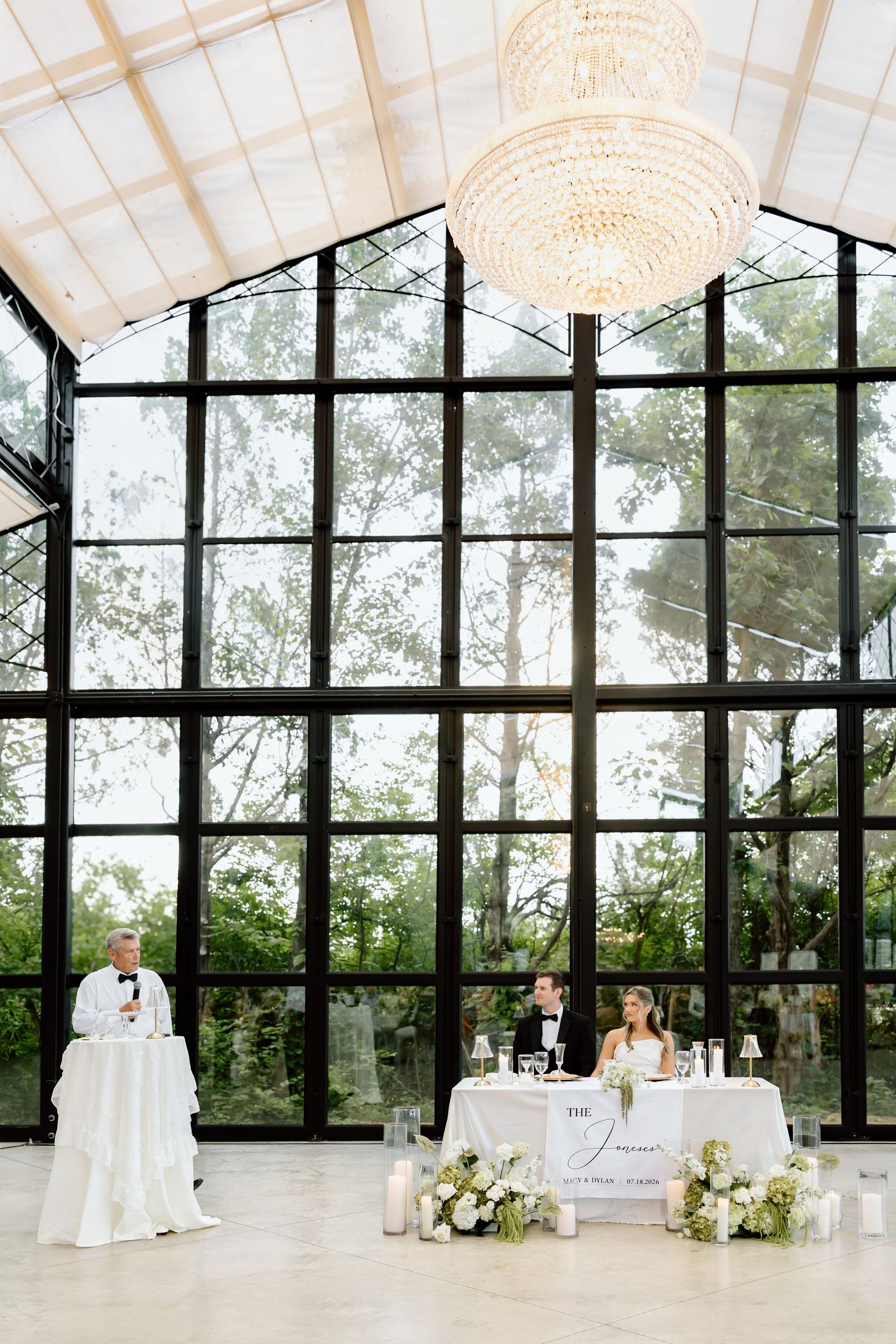 Elegant wedding reception in a glass greenhouse venue with chandelier, sweetheart table, and floral arrangements.