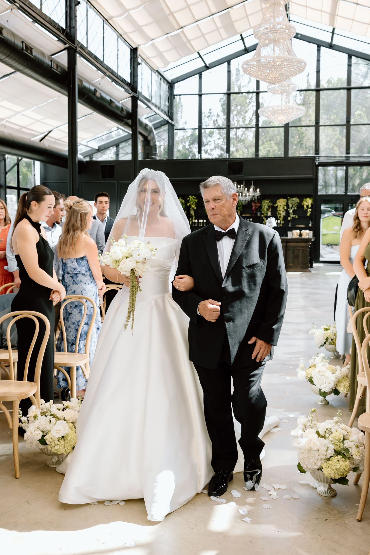 Bride in white gown and veil walks down aisle with older man in tuxedo in elegant glass greenhouse venue.