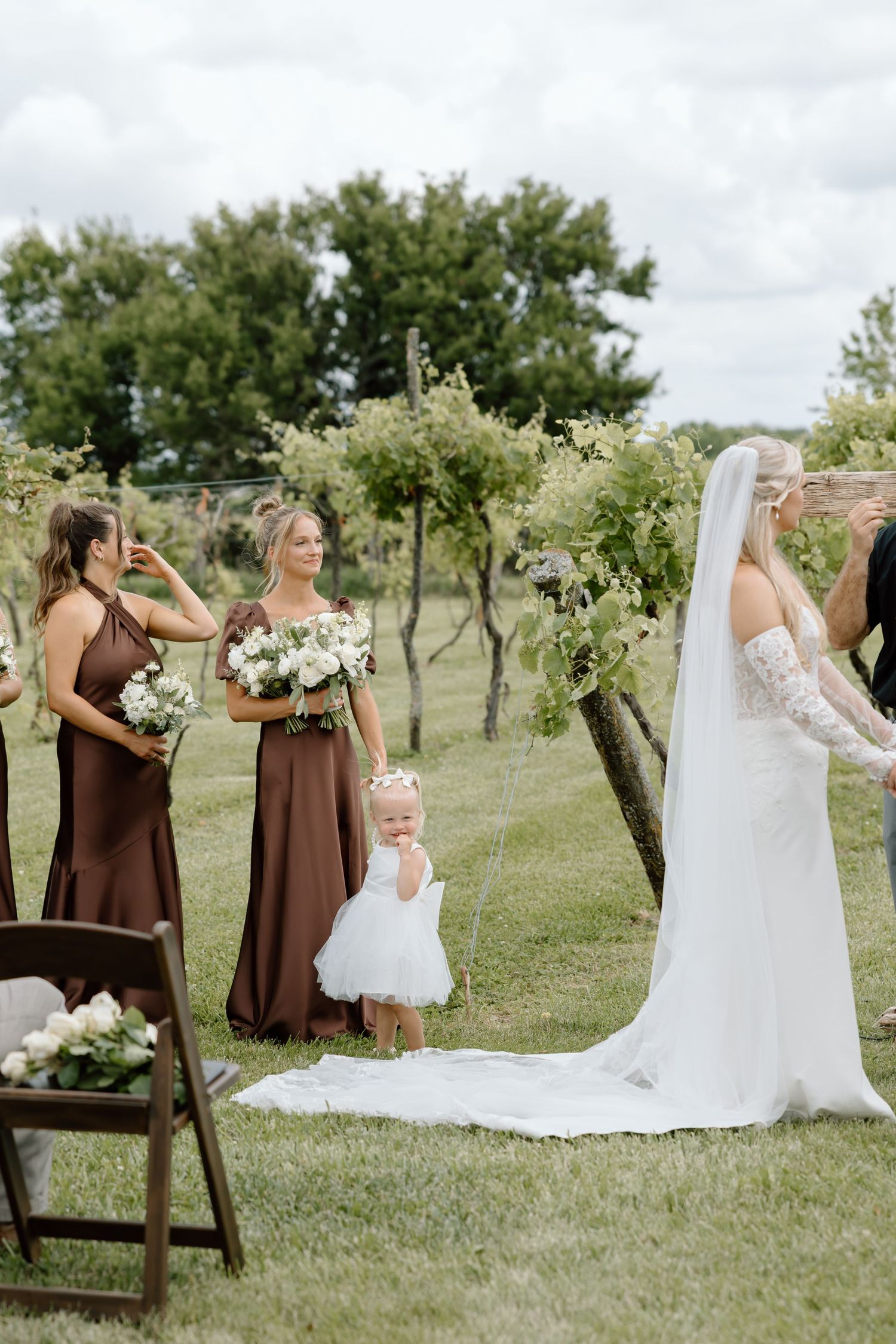 Bride in white gown with veil stands outdoors at vineyard wedding with bridesmaids in brown dresses and flower girl.