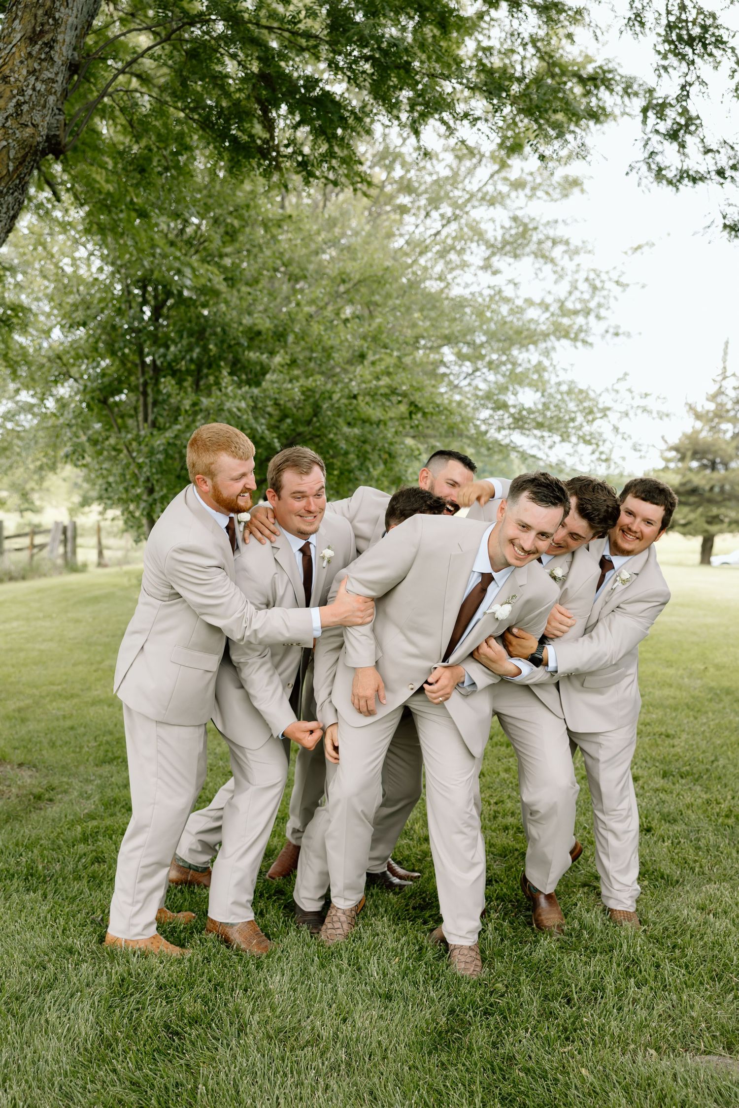 Groomsmen in light gray suits playfully hugging the groom outdoors on a green lawn at a wedding.