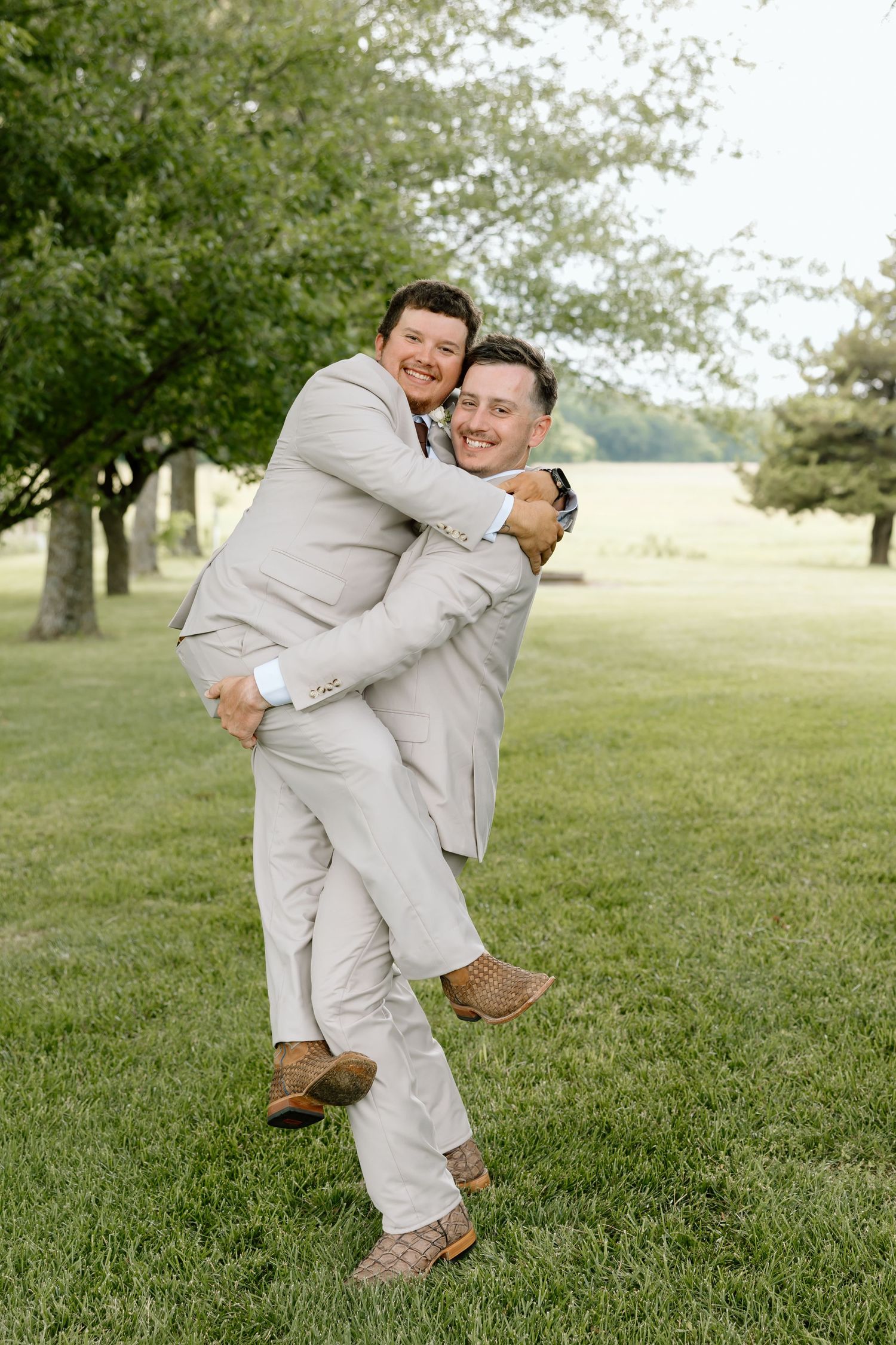 Two grooms in matching tan suits embrace joyfully outdoors on green grass with trees in the background.
