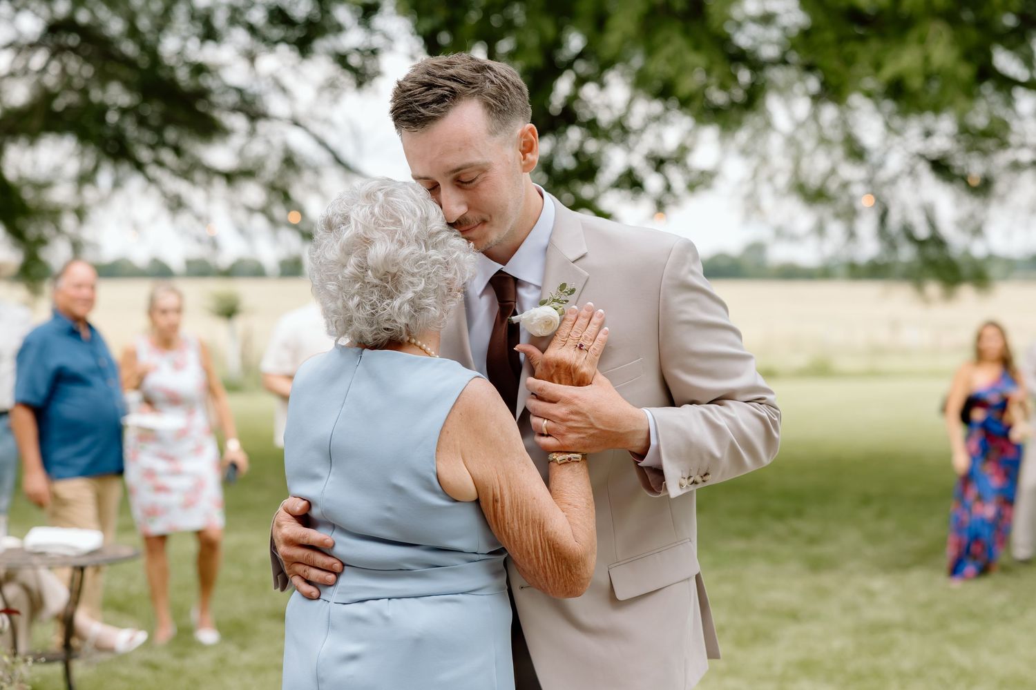 Groom in tan suit shares a tender dance with elderly woman outdoors at a wedding reception on a sunny day.