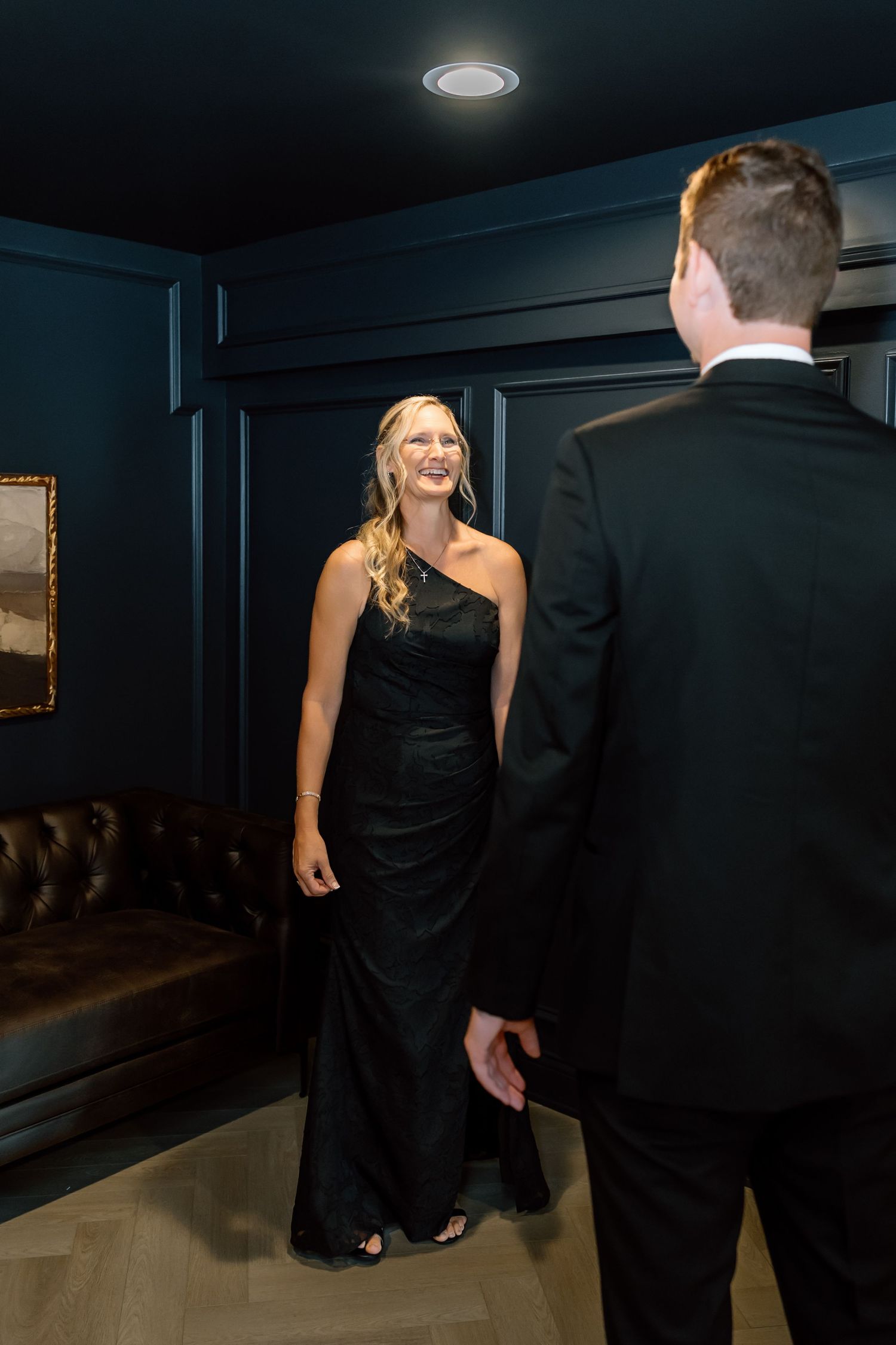 Woman in black one-shoulder gown smiling at man in tuxedo in elegant dark blue room.