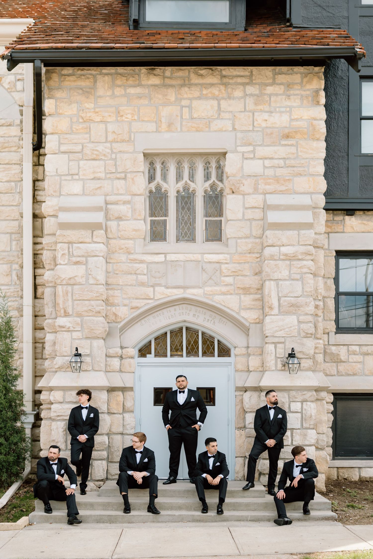 Groom and groomsmen in black tuxedos posing on steps of elegant stone building with Gothic arch doorway.