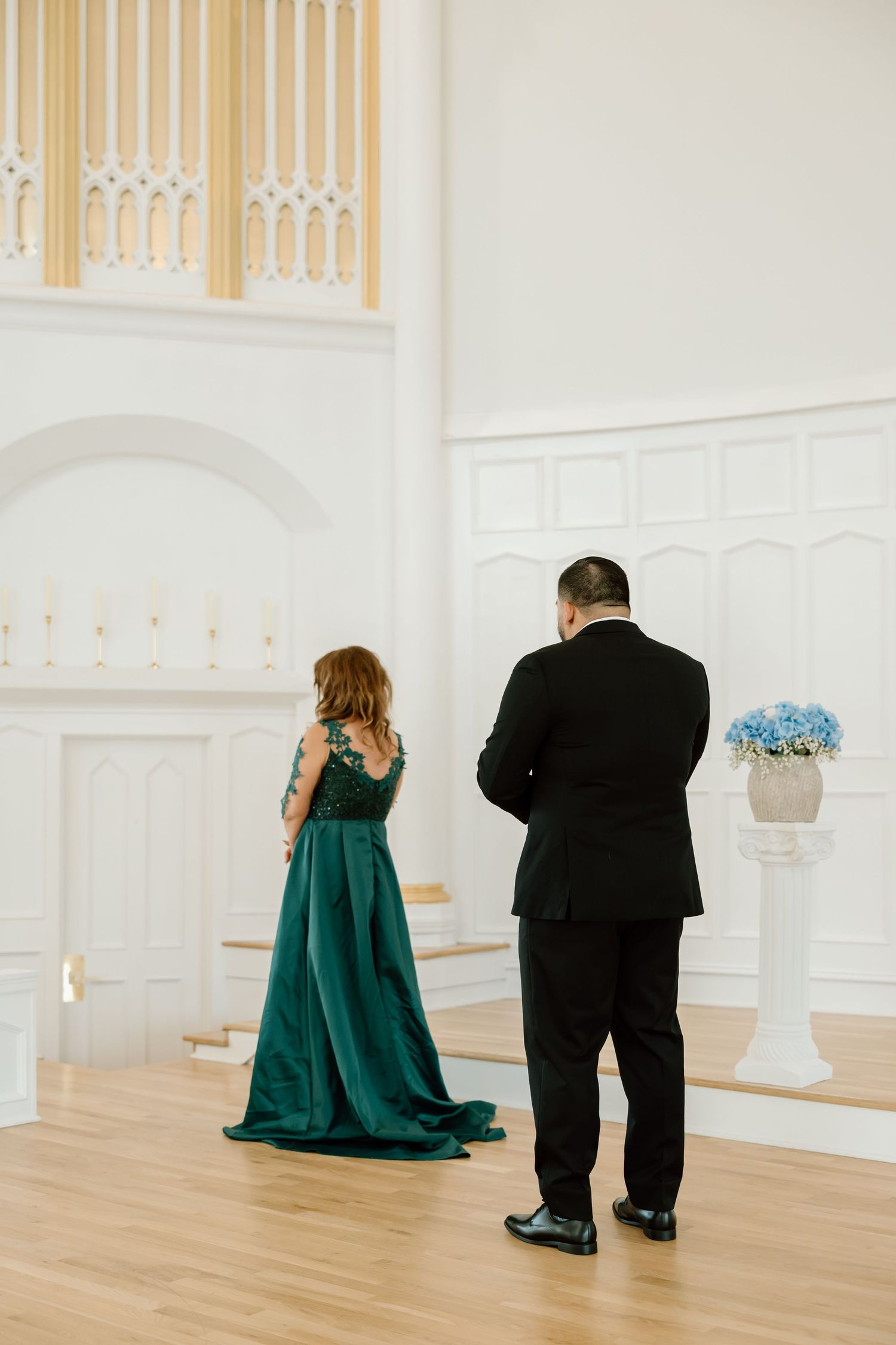 Couple in elegant attire, woman in green gown and man in black suit, standing in a white wedding venue.