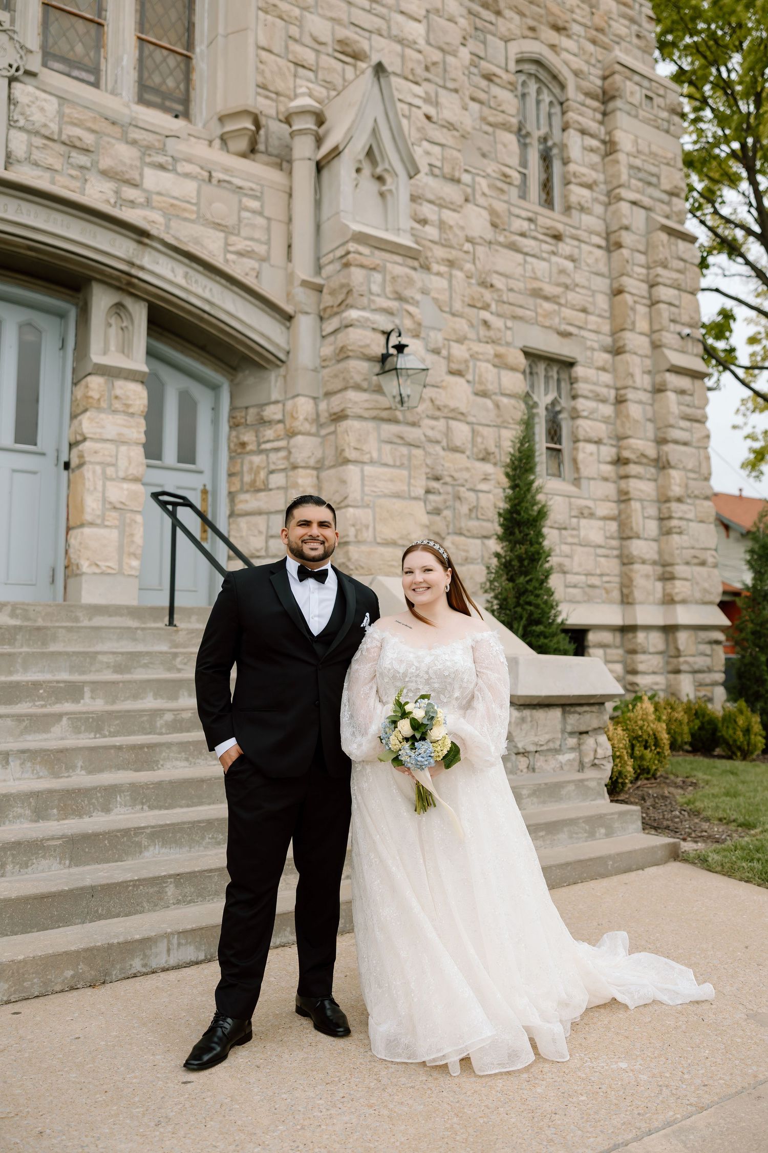 Bride and groom posing in front of a stone church, bride in a white lace gown holding a blue bouquet.