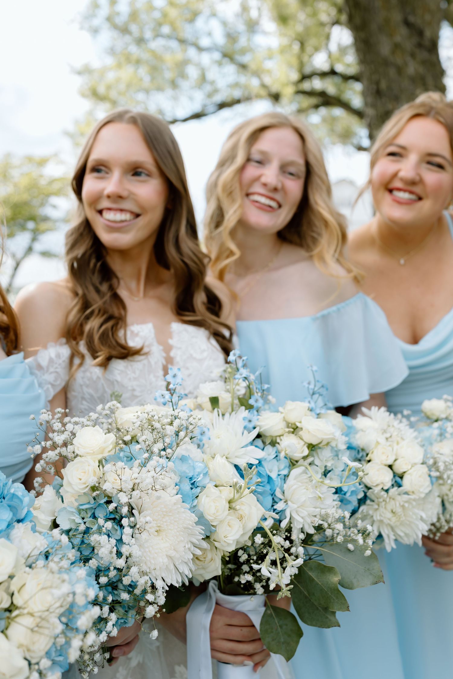 Bridesmaids in light blue off-shoulder dresses holding white and blue floral bouquets, smiling outdoors at a wedding.