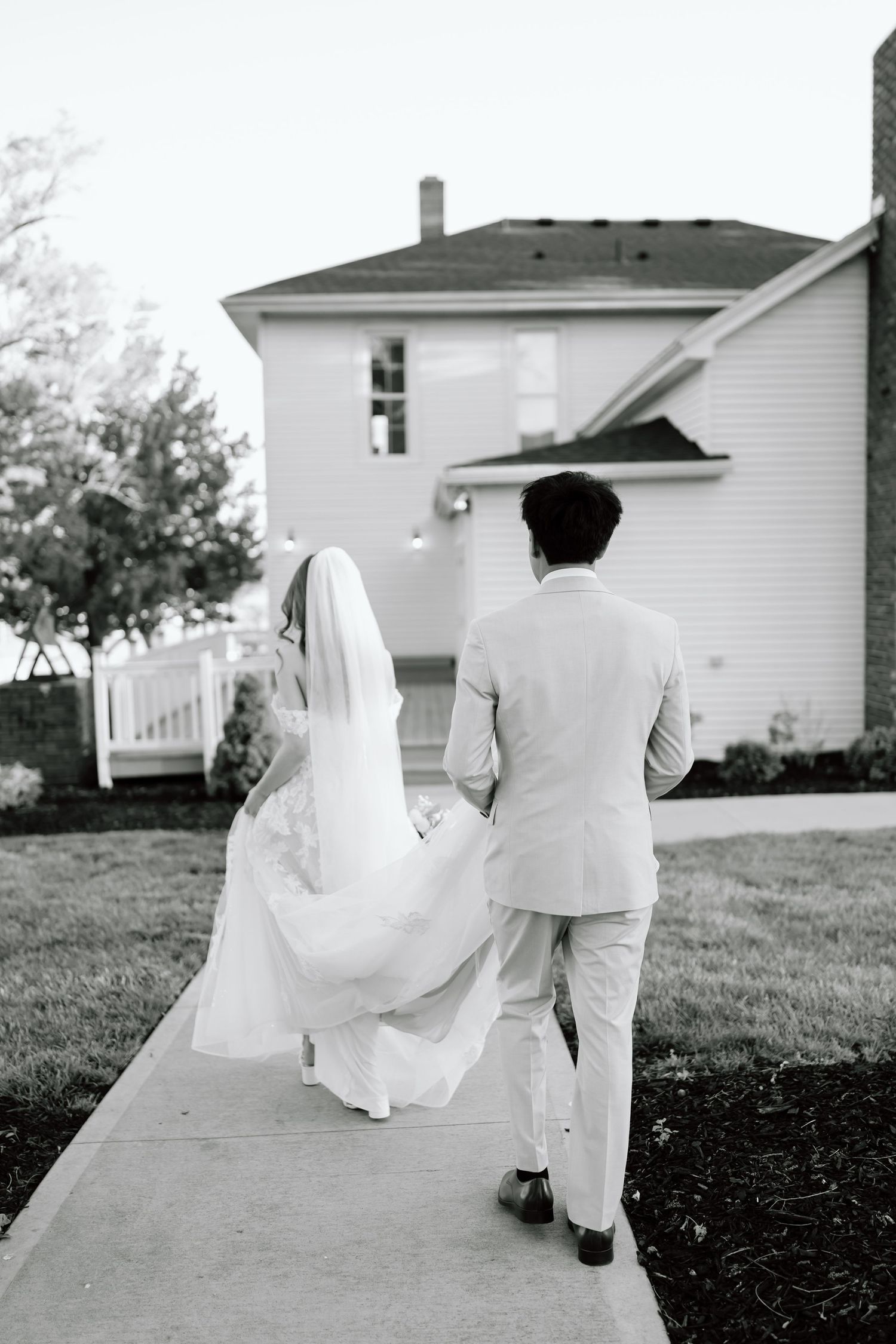 Black and white photo of a bride and groom walking hand in hand down a pathway toward a house.