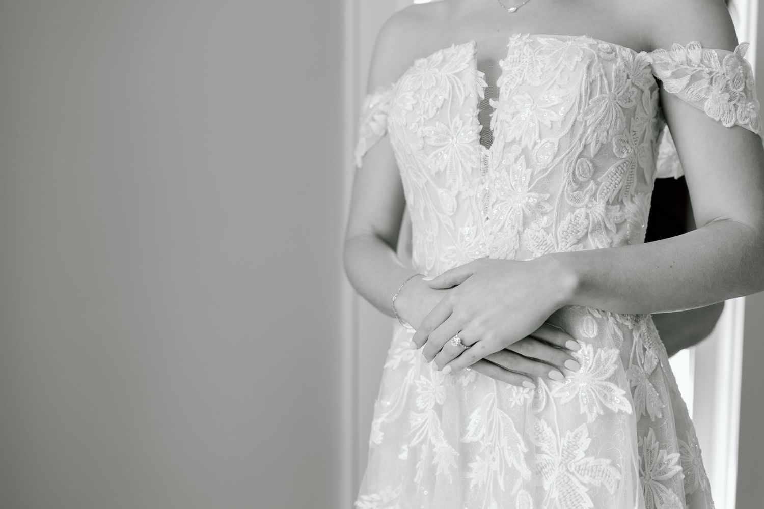 Black and white photo of a bride in an off-shoulder lace wedding gown, hands folded gracefully at her waist.