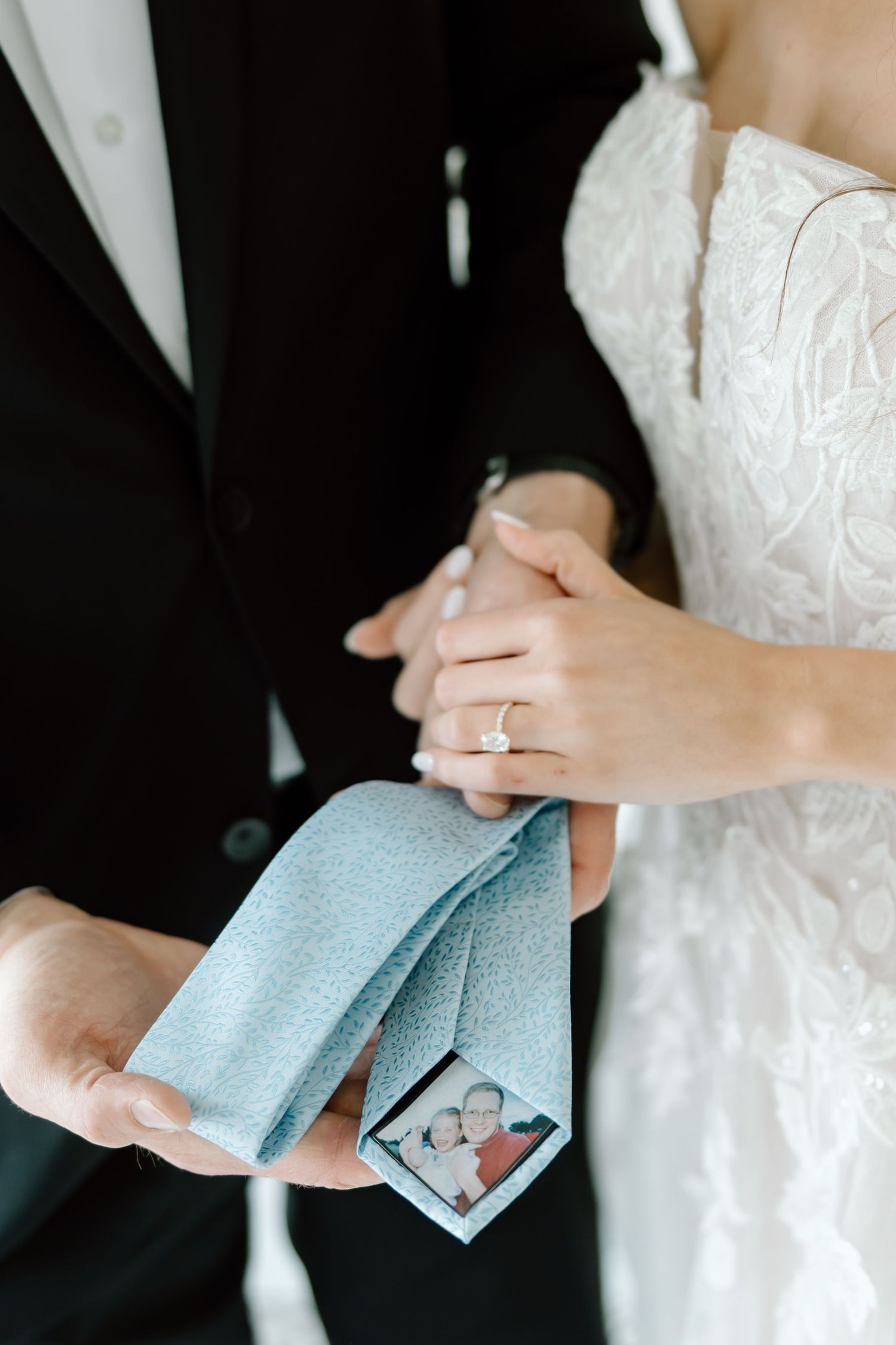 Bride and groom holding a light blue tie with a small photo patch, bride wearing an engagement ring and lace wedding dress.