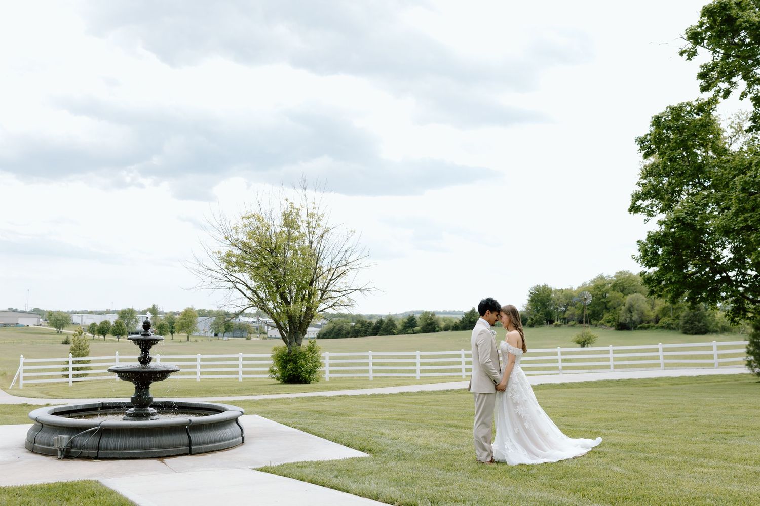 A bride and groom share a romantic moment on a lush green lawn beside a stone fountain at an elegant outdoor wedding venue.