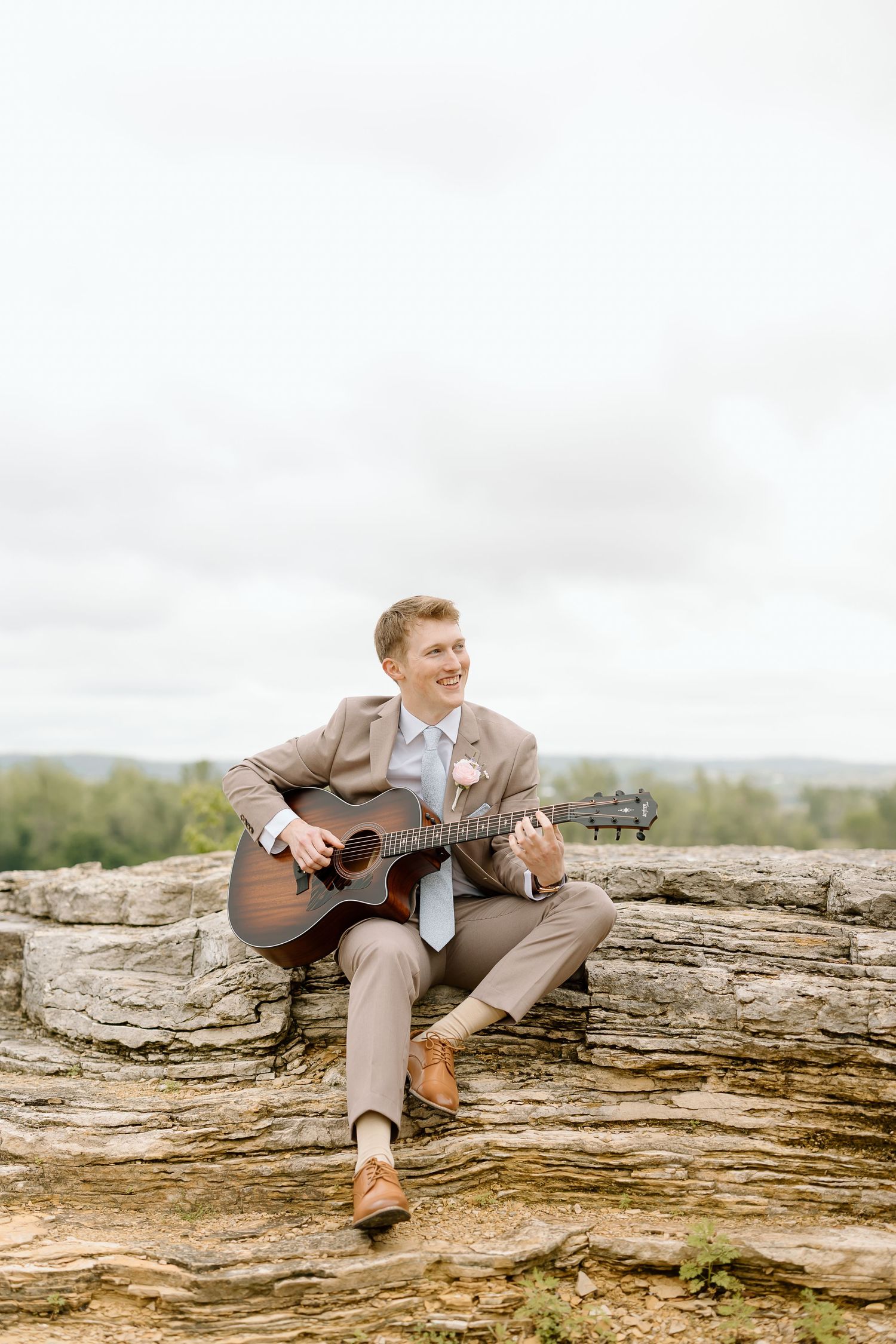 A person in a gray suit plays acoustic guitar while sitting on a large fallen tree trunk in a natural outdoor setting.